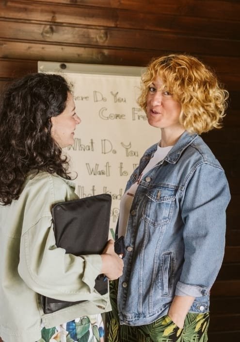 Two women having a conversation indoors, with a wooden wall and a whiteboard in the background.