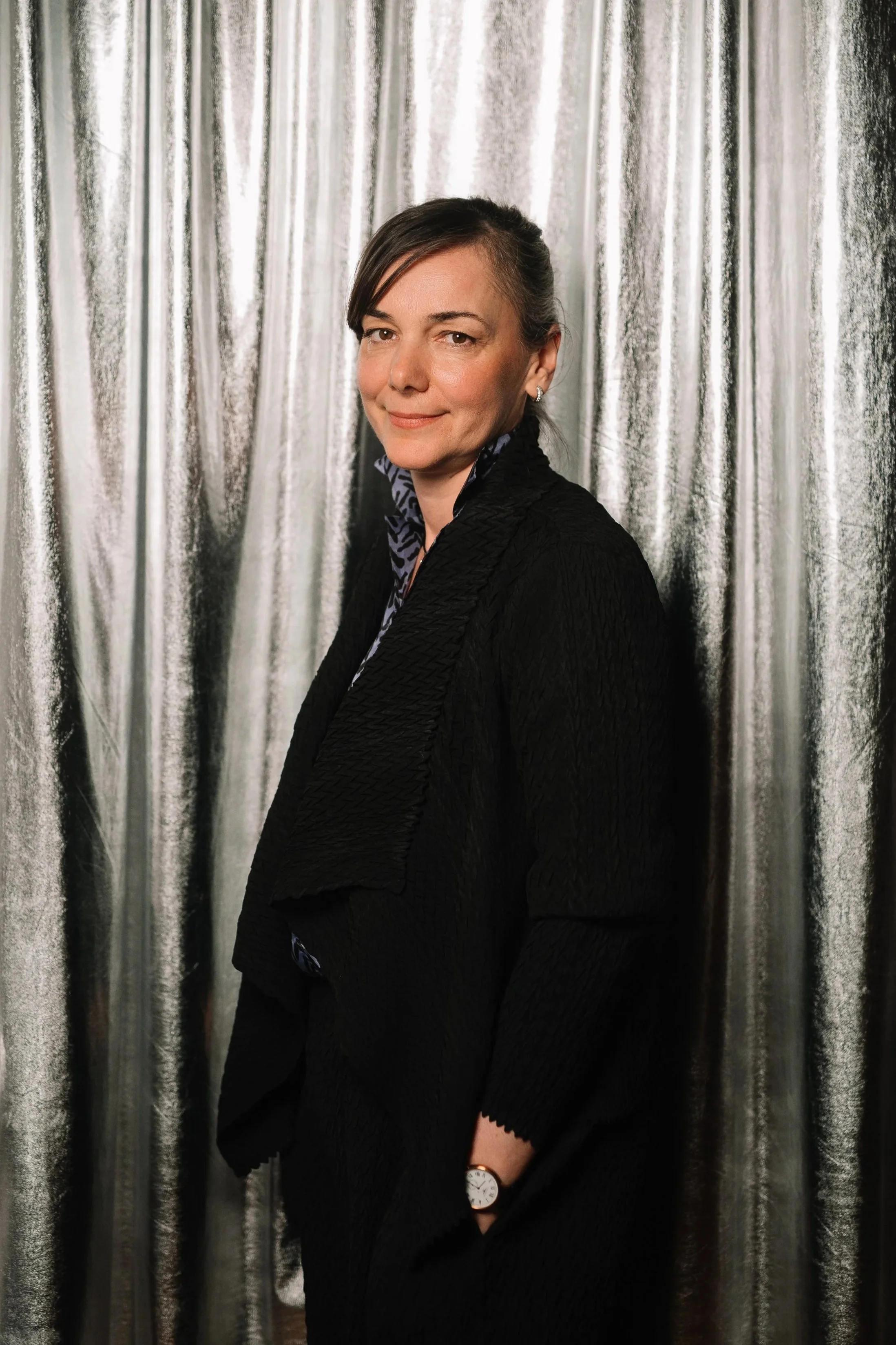 A woman with dark hair styled in a side part, wearing a black textured blazer over a patterned shirt, standing in front of a silver curtain backdrop, smiling slightly.