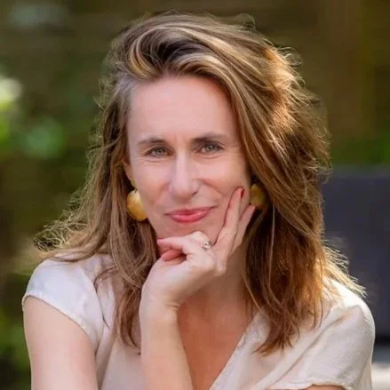 A woman with shoulder-length wavy hair, wearing a white top and yellow earrings, resting her chin on her hand and smiling at the camera outdoors.