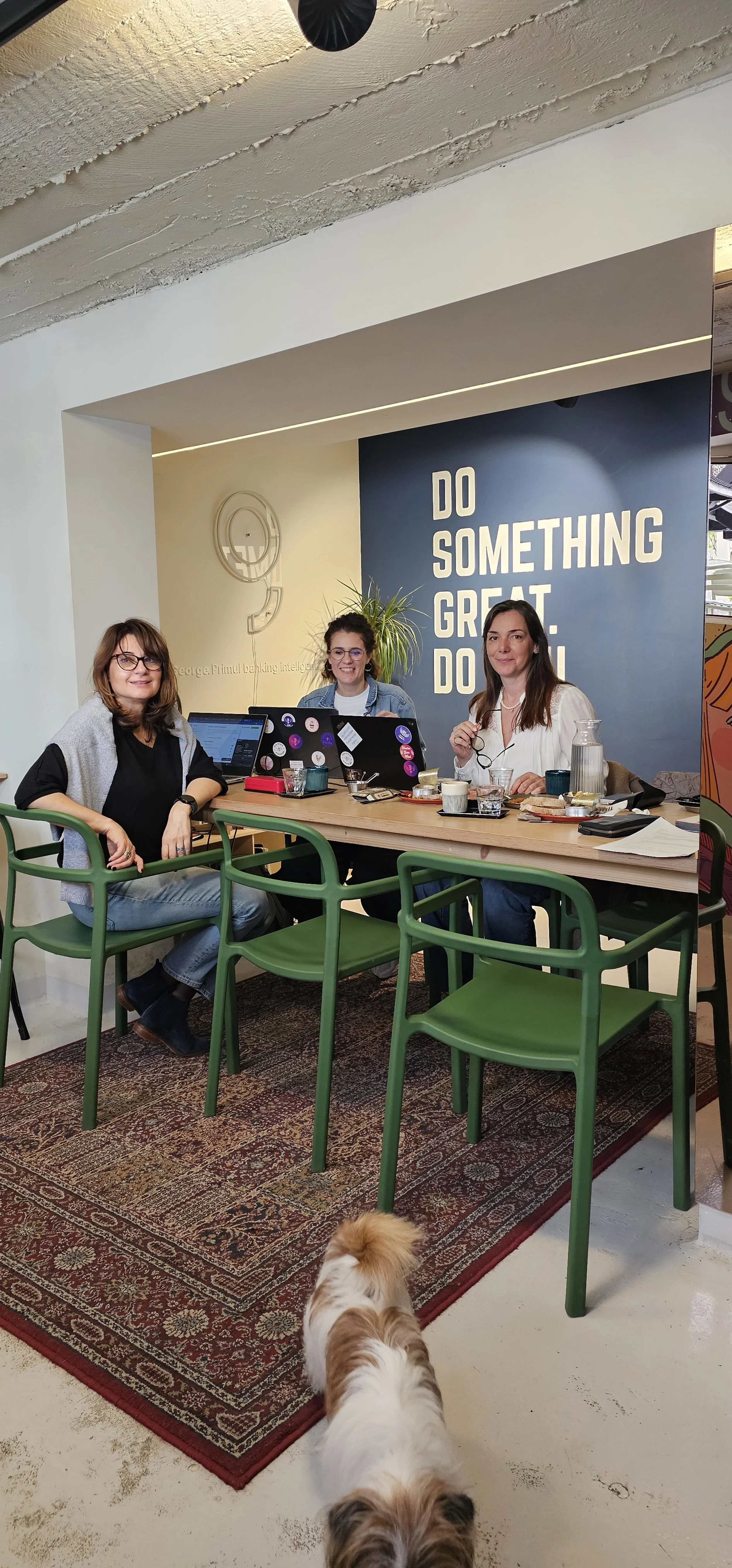 Three women sitting at a wooden table in a modern cafe with a patterned rug. One woman with glasses and brown hair wearing a black shirt and gray vest, another with glasses and curly hair, and the third with long brown hair in a white shirt. There are laptops, plates, cups, and a glass pitcher on the table. A small dog with white and brown fur is walking in front. The background has a dark blue wall with white text that reads 'Do Something Great. Do.'