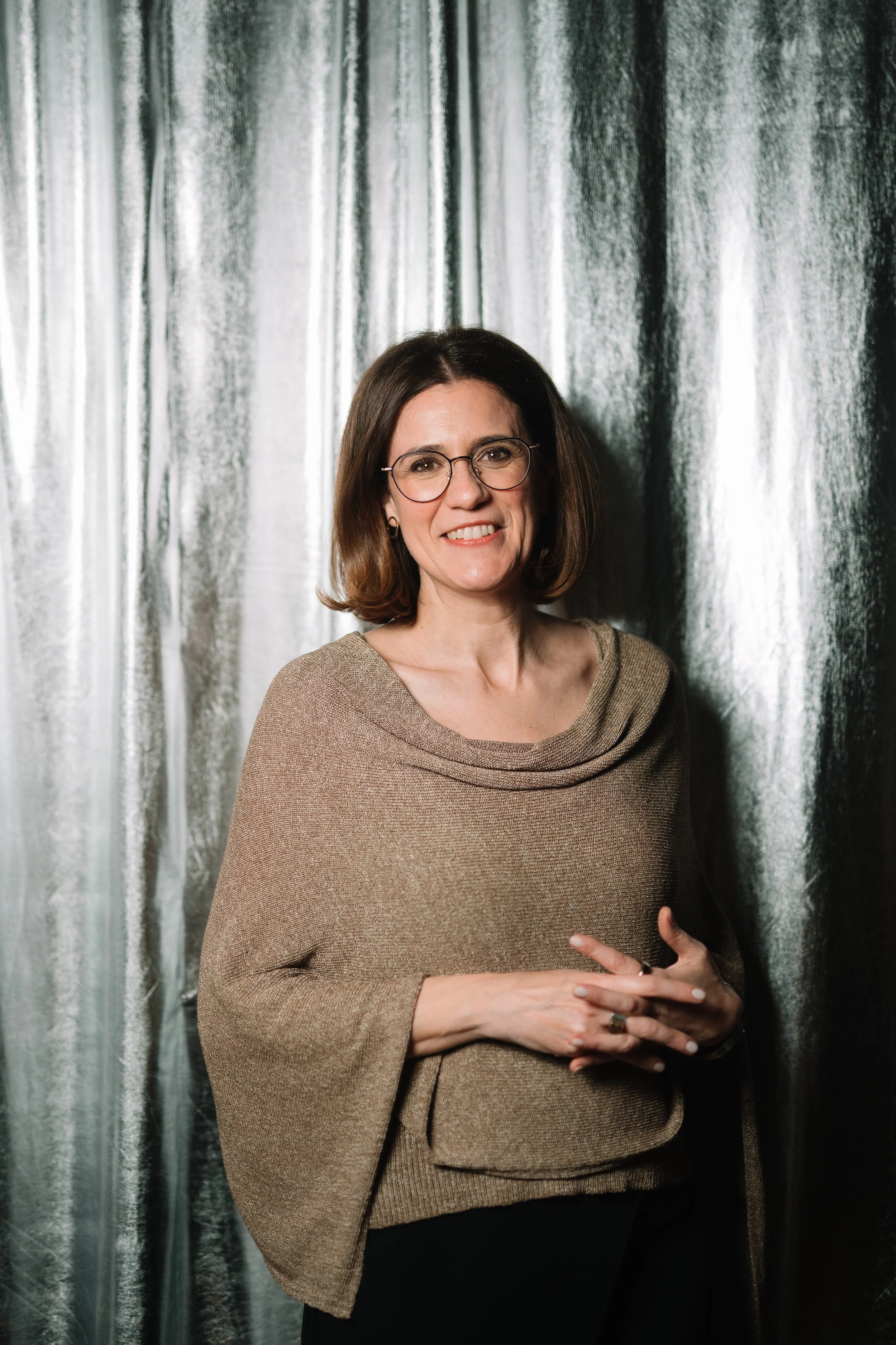 A woman with short brown hair, glasses, wearing a tan knitted top, standing in front of a shiny silver curtain.
