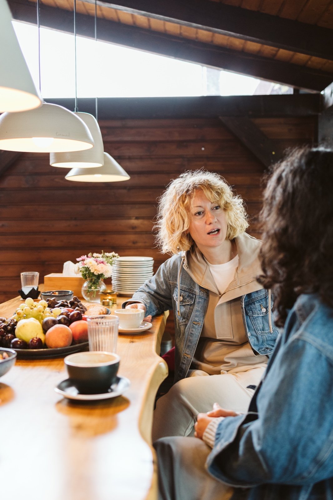 Two women sitting at a wooden table in a cozy, wood-paneled room, engaged in conversation. The table is set with bowls, a plate of assorted fruits, cups, and a small flower arrangement. One woman has curly blonde hair, wearing a denim jacket, and appears to be speaking.