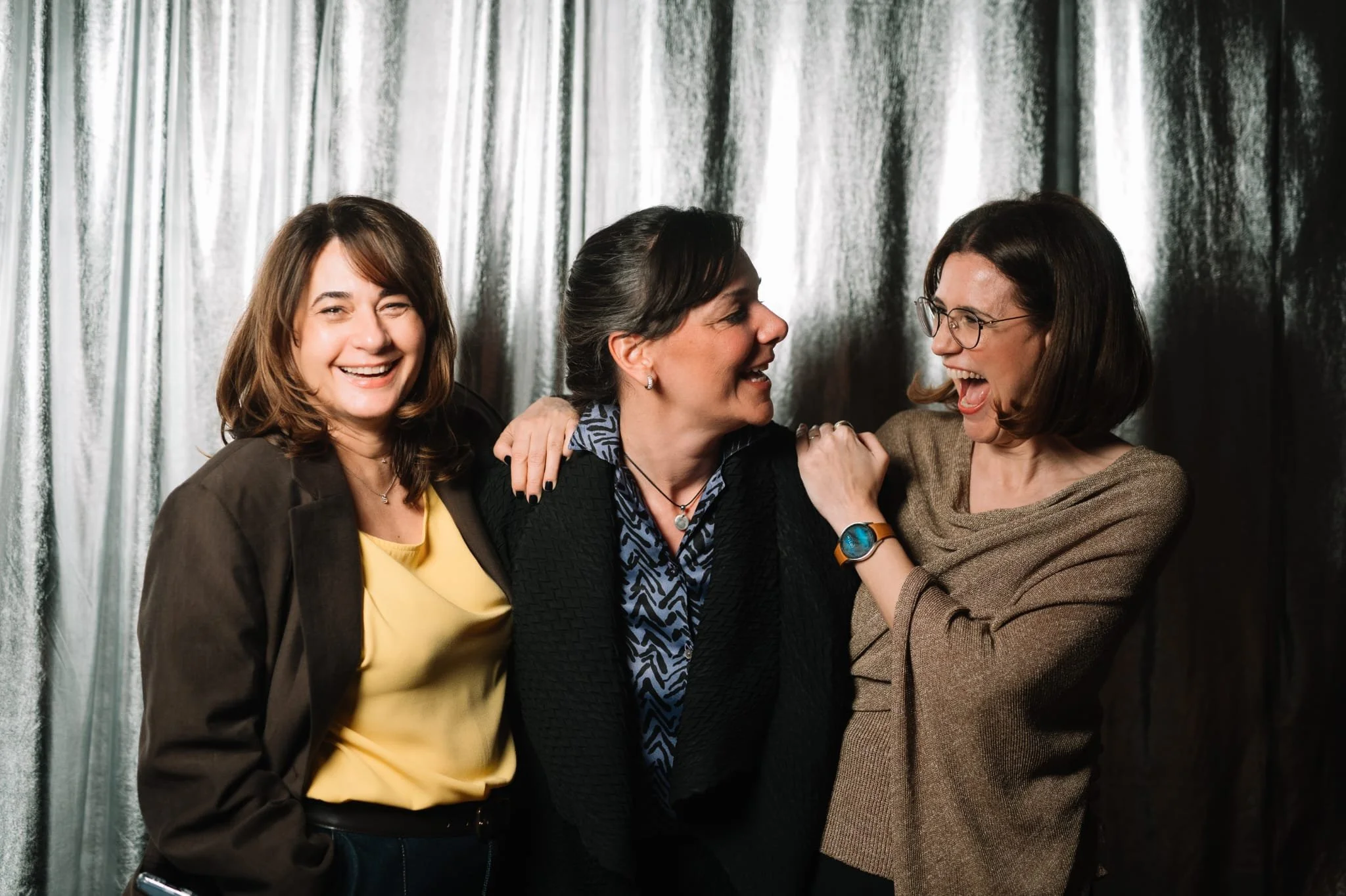 Three women smiling and laughing together in front of a silver curtain, with two women on each side and one in the middle.
