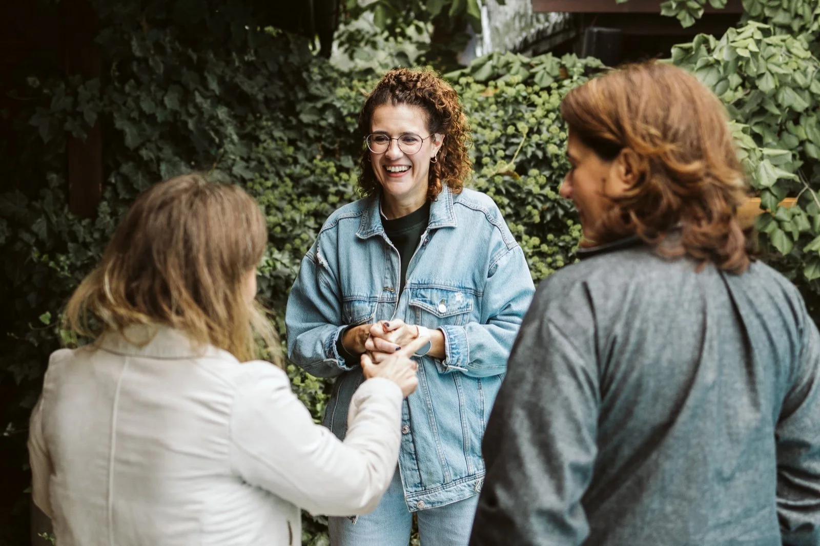 Three women outdoors, one woman with curly hair and glasses smiling, holding hands with a blonde woman in a white jacket, while another woman with straight hair and a gray jacket looks on.