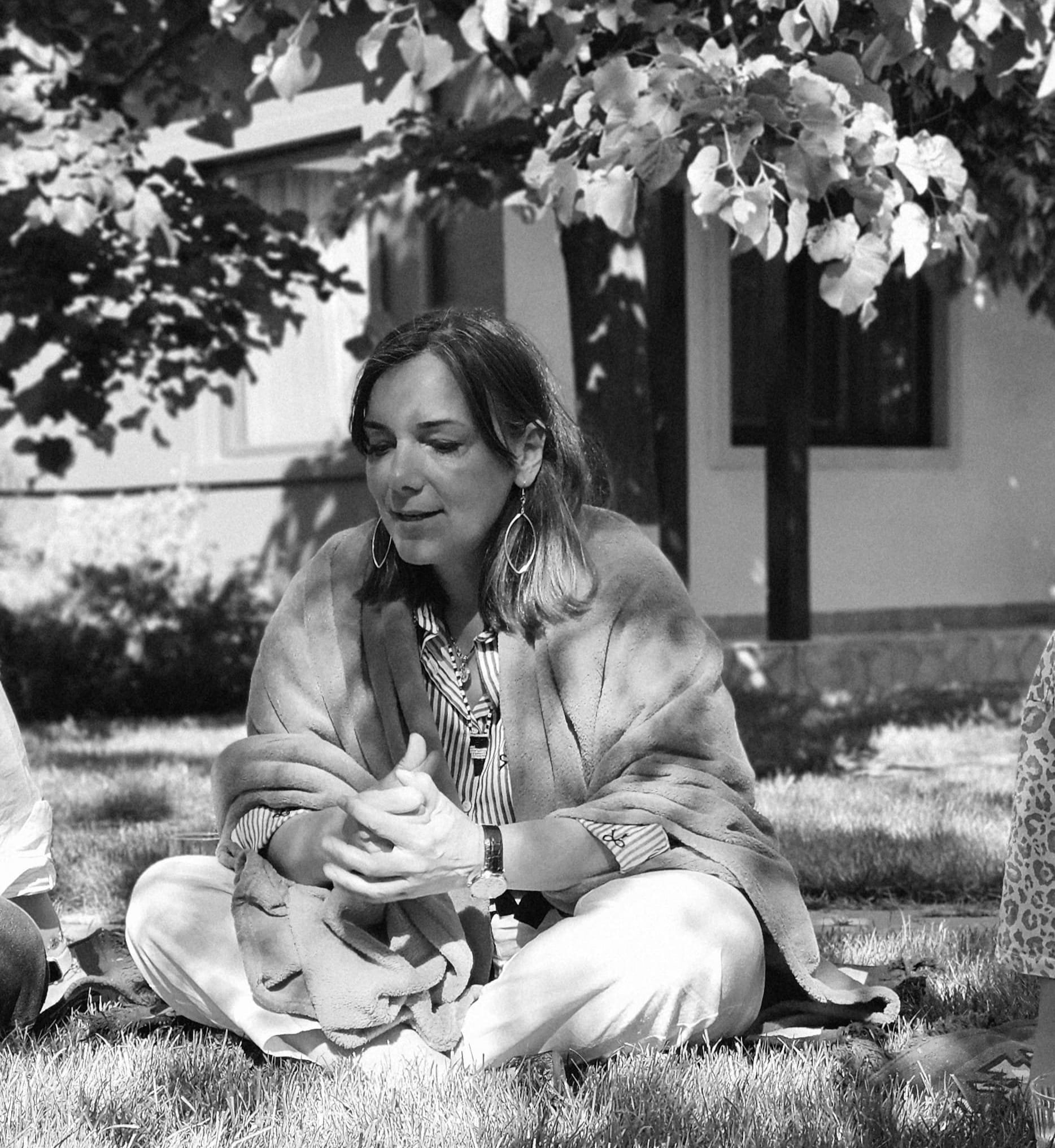 A woman sitting on the grass, wrapped in a blanket, outdoors at night with trees and a house in the background.