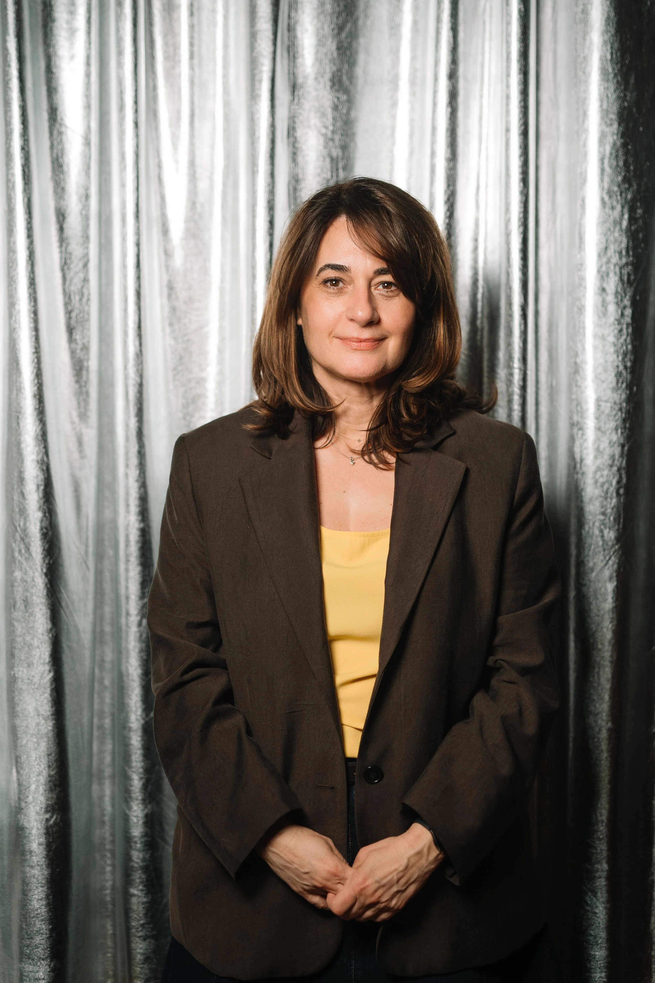 A woman with shoulder-length brown hair, wearing a brown blazer and yellow top, standing in front of a silver curtain backdrop, smiling softly.