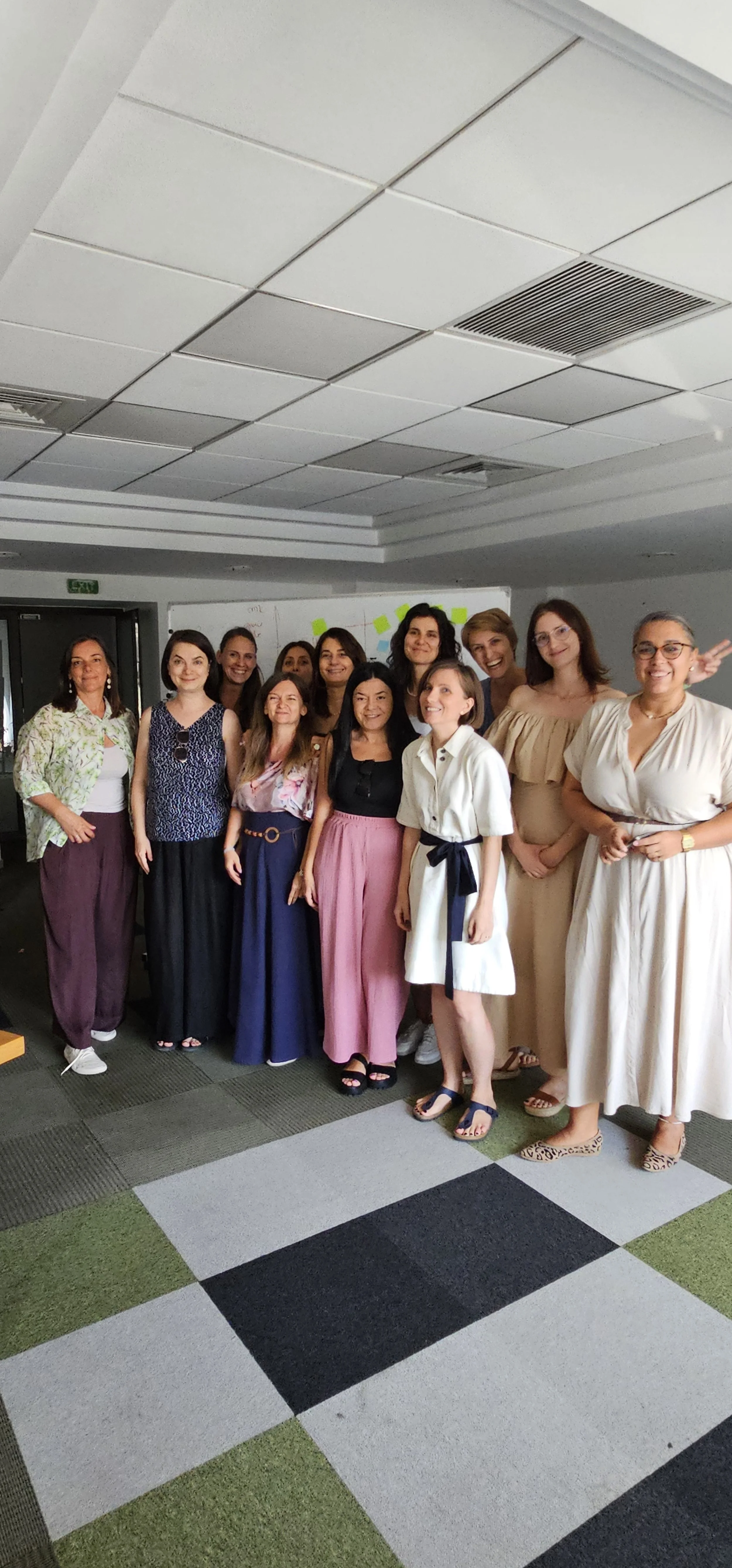 Group of women standing together indoors, smiling for a photo, with a whiteboard behind them and a ceiling with vents.