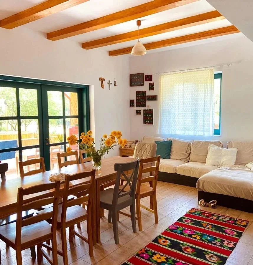 A cozy dining and living room area with wooden beams on the ceiling, a large window with white curtains, a sliding door with green trim leading outside, a wooden dining table with chairs and a vase of yellow flowers, a white sectional sofa with throw pillows, a colorful floral rug, and wall decorations including framed artwork and small crosses.