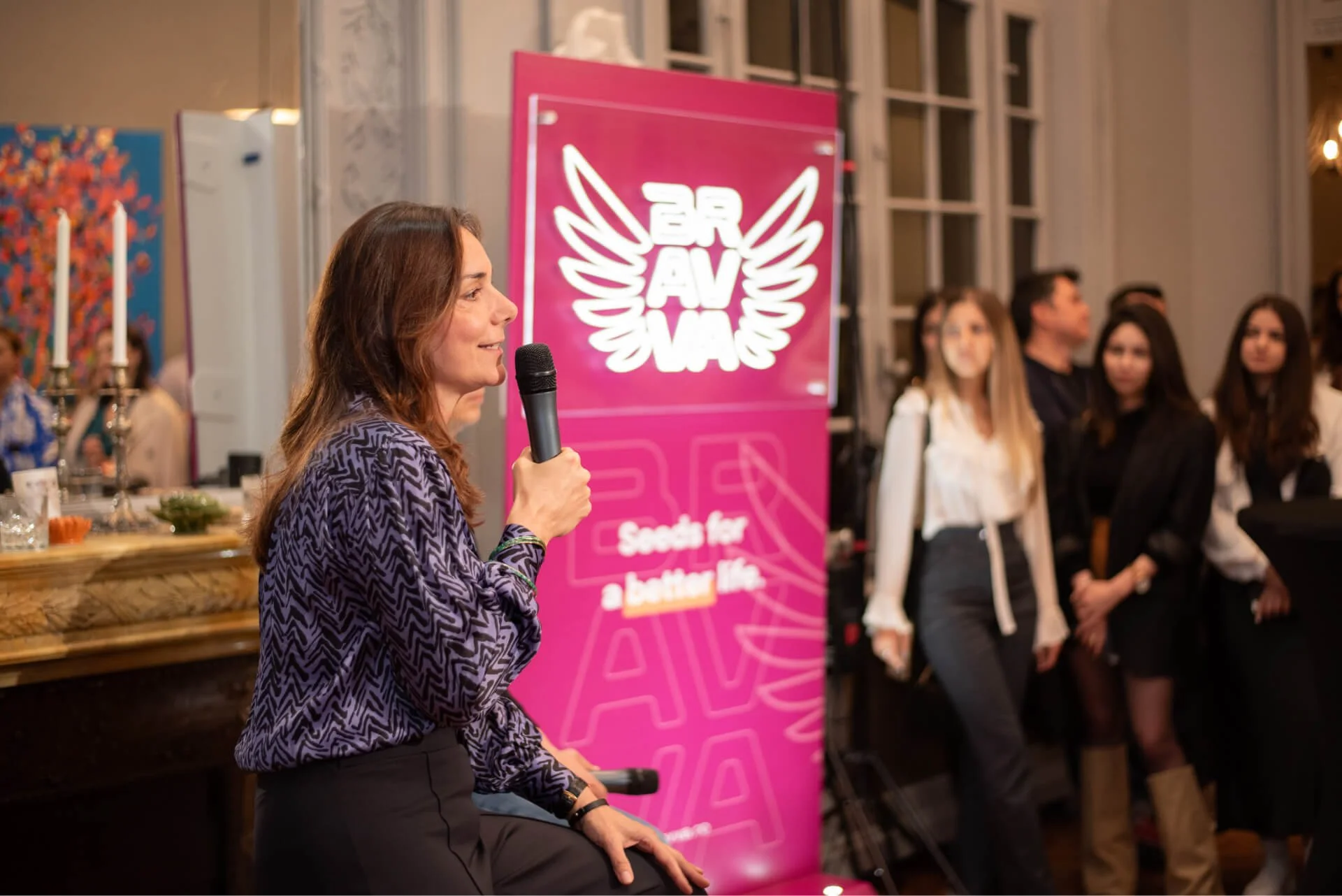 A woman holding a microphone speaking at an event with a pink sign behind her that reads 'BRAVO' with wings and the slogan 'Seeds for a better life.' Several people stand in the background, listening.
