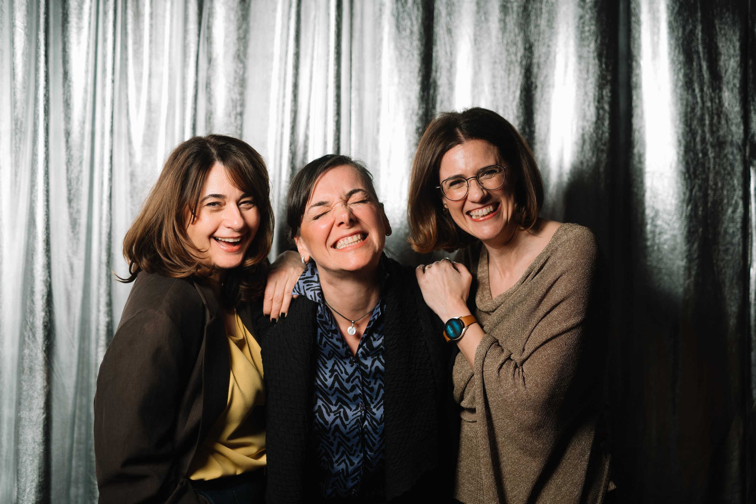 Three women smiling and hugging each other against a black curtain background.