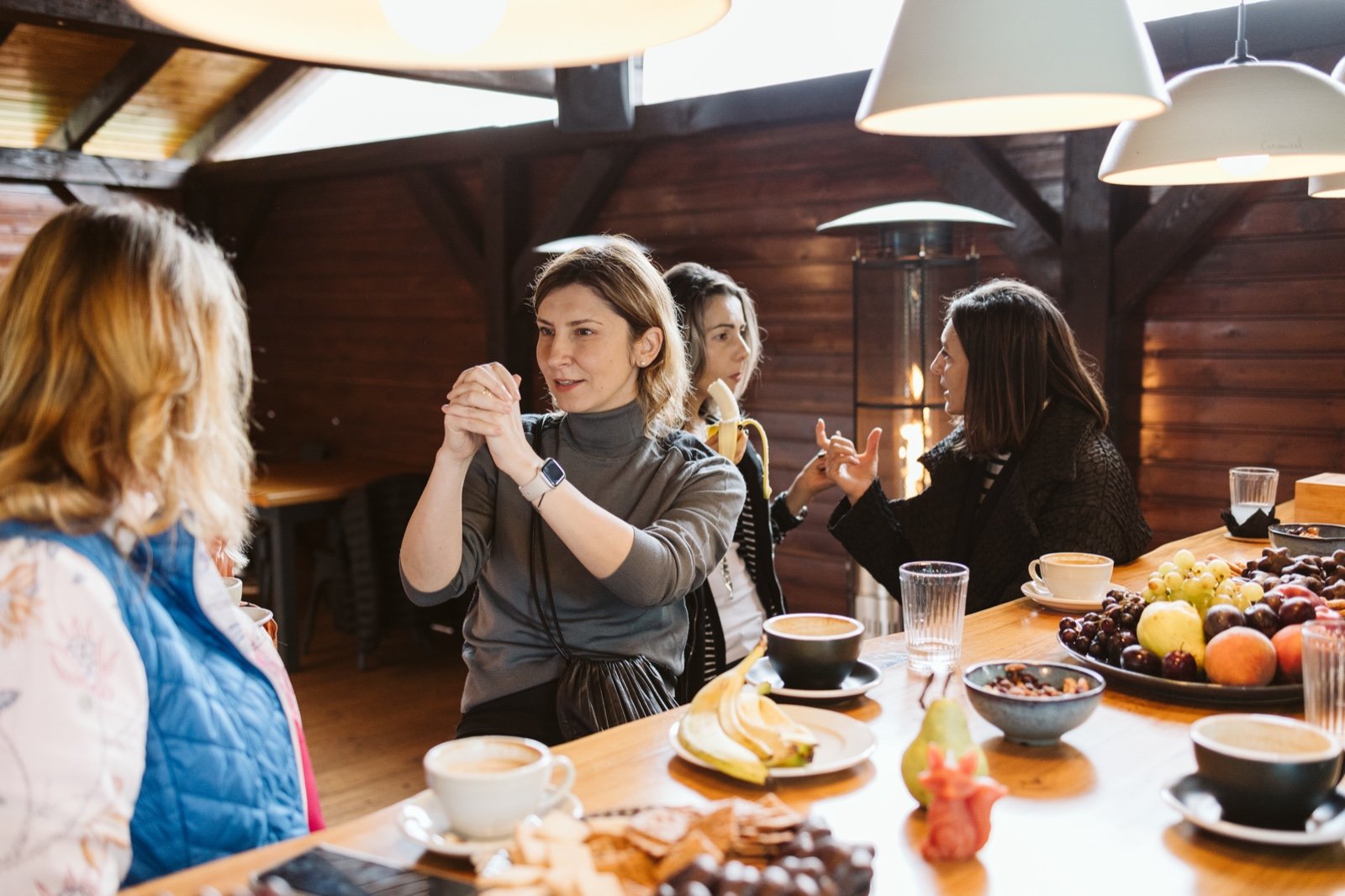 Four women sitting around a wooden table in a cozy, rustic interior, enjoying coffee and snacks, with plates of fruit and chocolates, engaged in conversation.