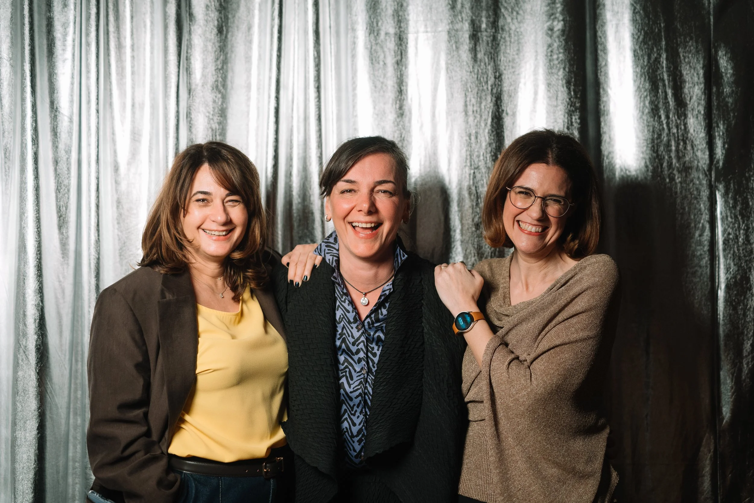 Three women smiling and laughing together in front of a shiny silver curtain.