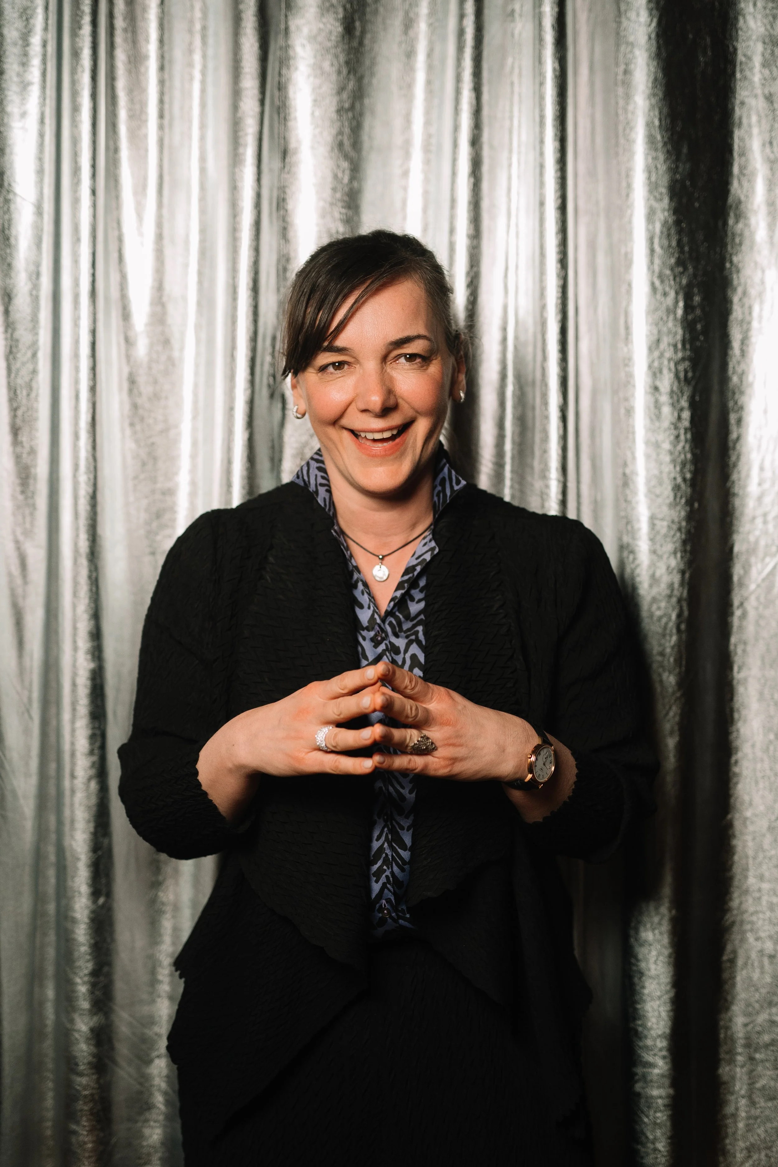A woman with short brown hair, smiling and wearing a black blazer, patterned blouse, and jewelry, standing in front of metallic silver curtains.