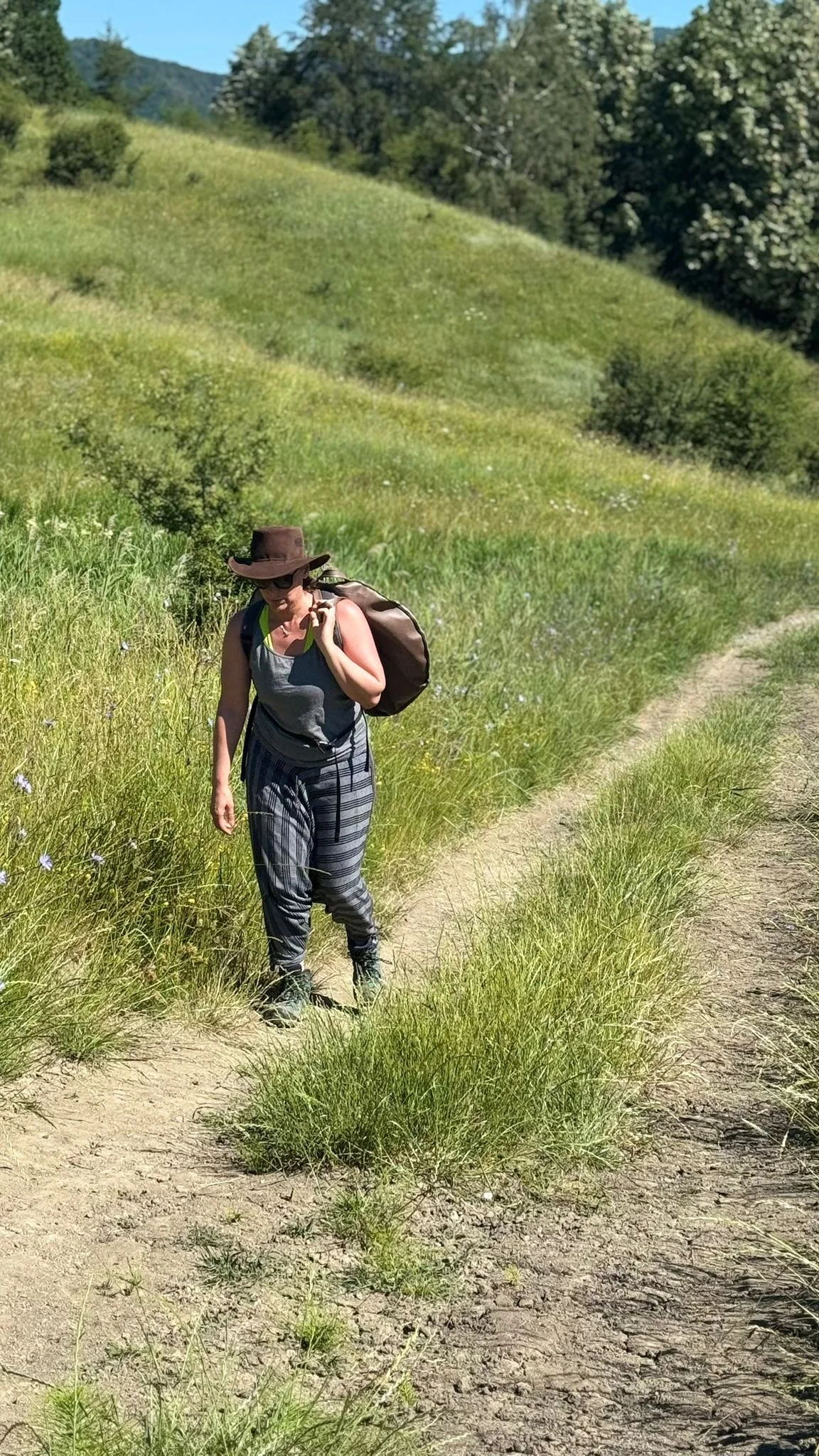 A woman hiking in a grassy landscape with hills and trees in the background, carrying a backpack and wearing a wide-brimmed hat and sunglasses.