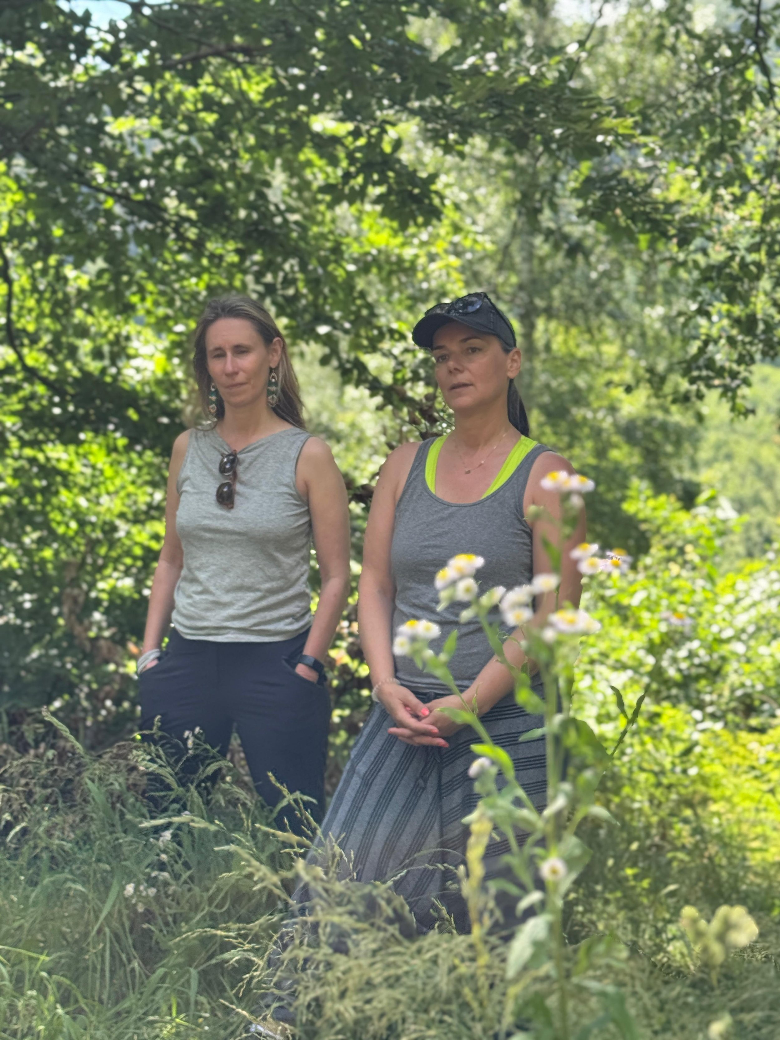 Two women standing outdoors in a lush green forest, one wearing a gray tank top with neon accents and a cap, the other wearing a sleeveless gray top with sunglasses hanging from her neckline.
