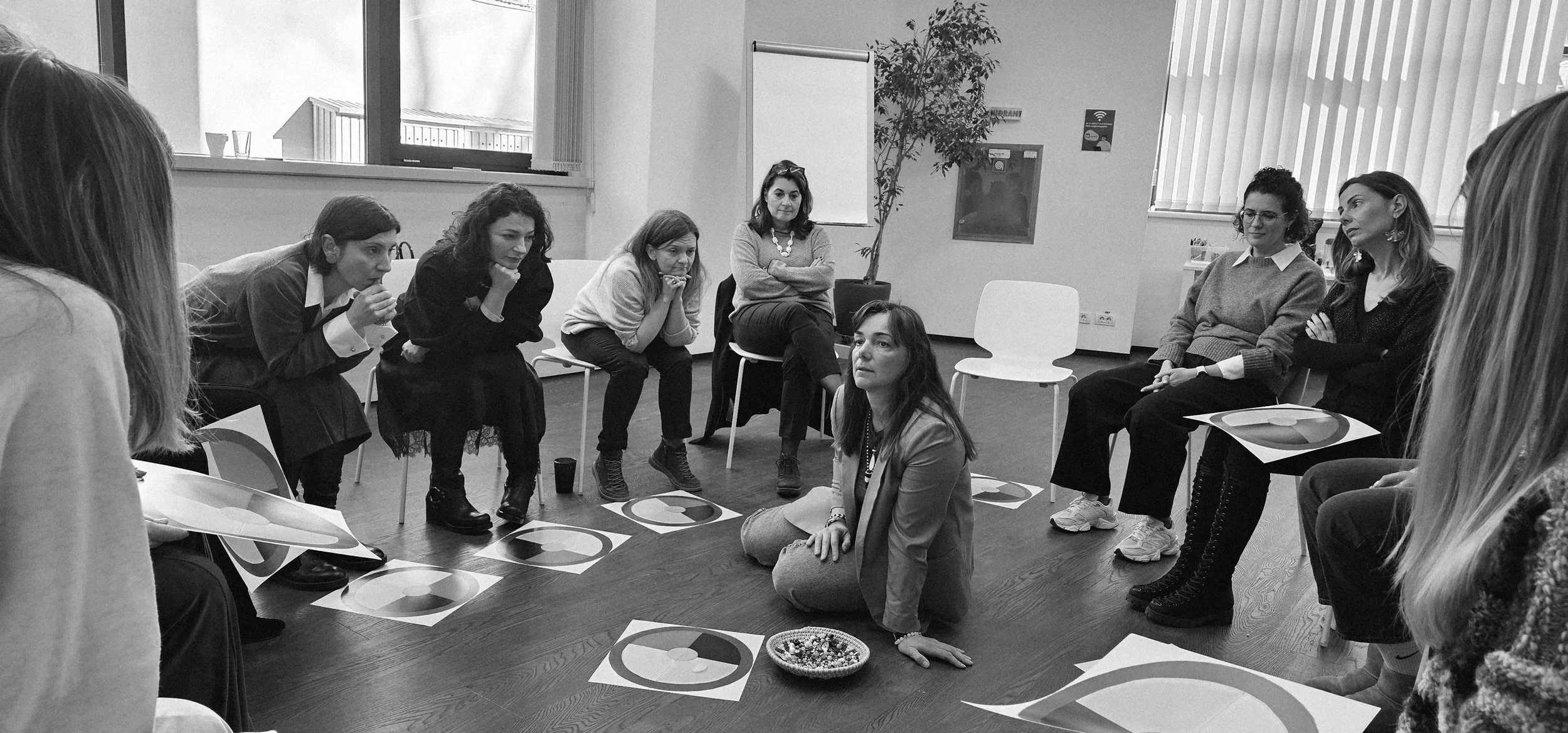 A group of women sitting in a circle in a room, engaging in a discussion or activity, with papers on the floor and a woman kneeling in the center.