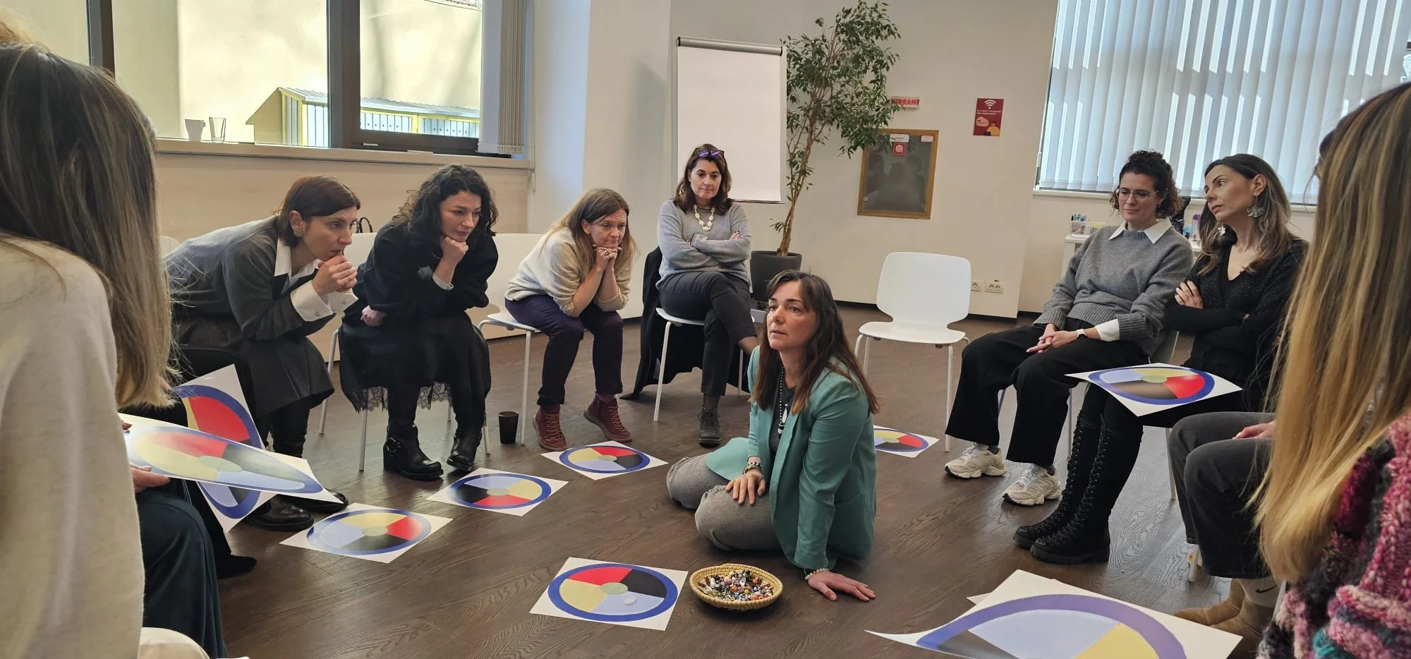 Group of women sitting and listening to a woman sitting on the floor with a basket of beads, in a circle indoors with colorful circular charts on the floor.