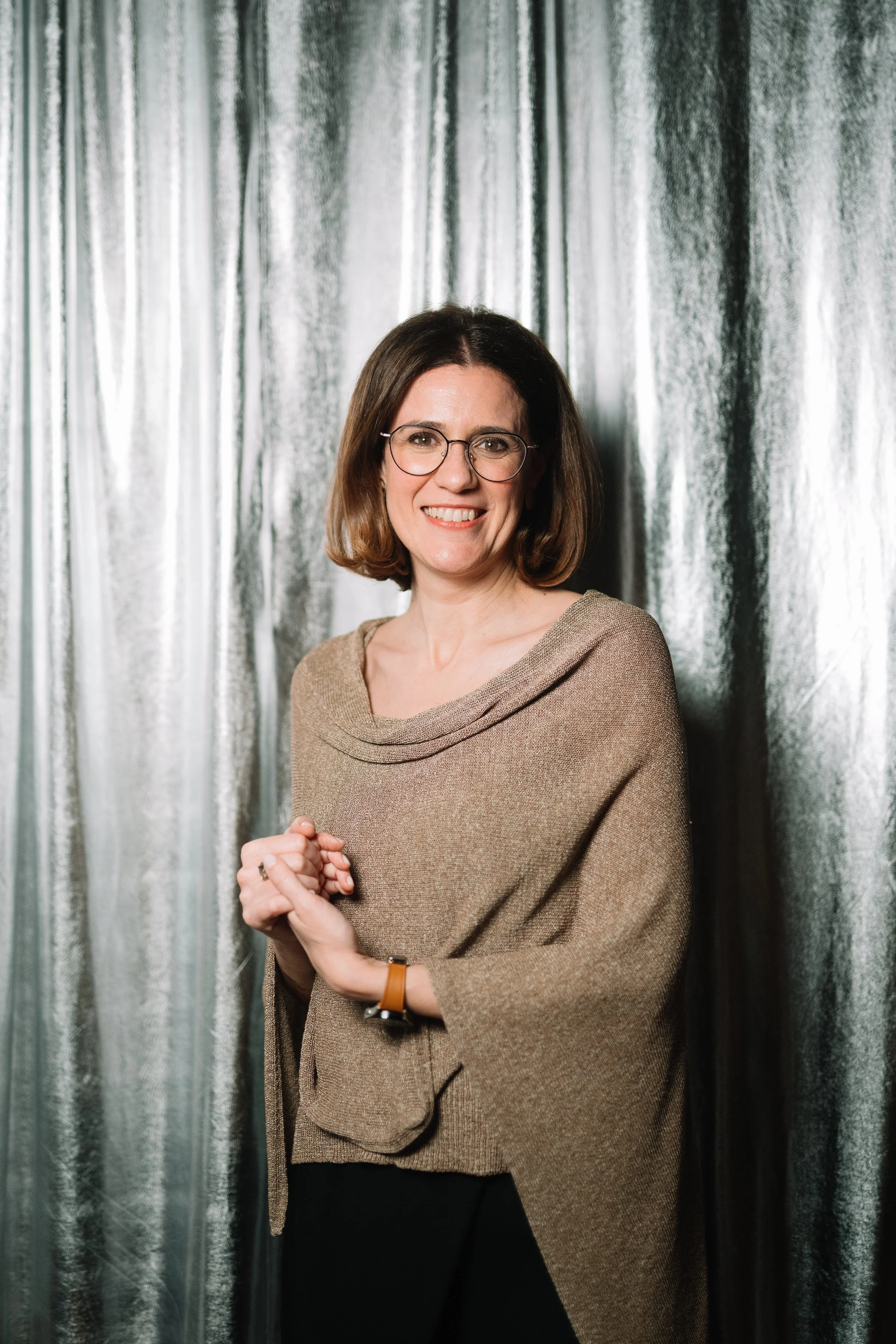 A woman with shoulder-length brown hair, glasses, and a beige sweater, standing in front of a silver metallic curtain and smiling at the camera.