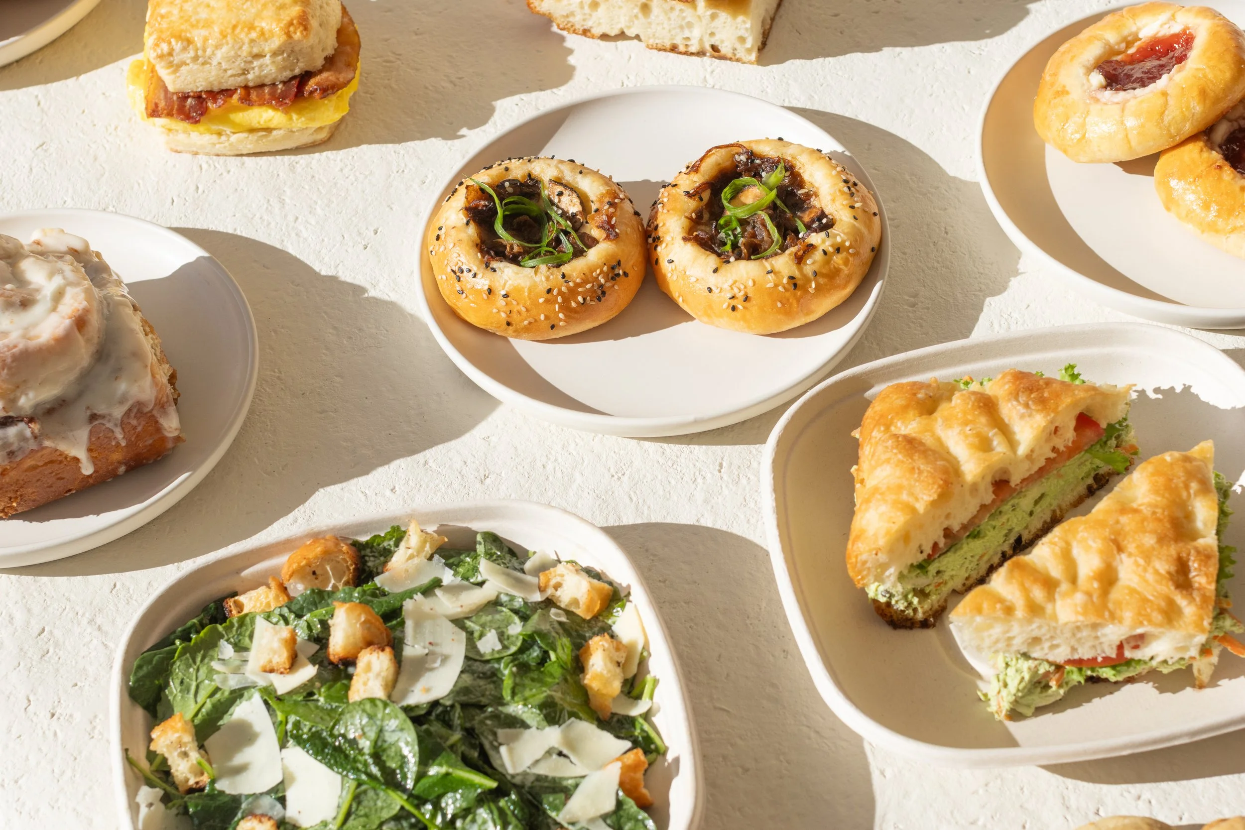 Assorted breakfast foods on white plates, including salad, sandwiches, bagels, and pastries with fruit filling.