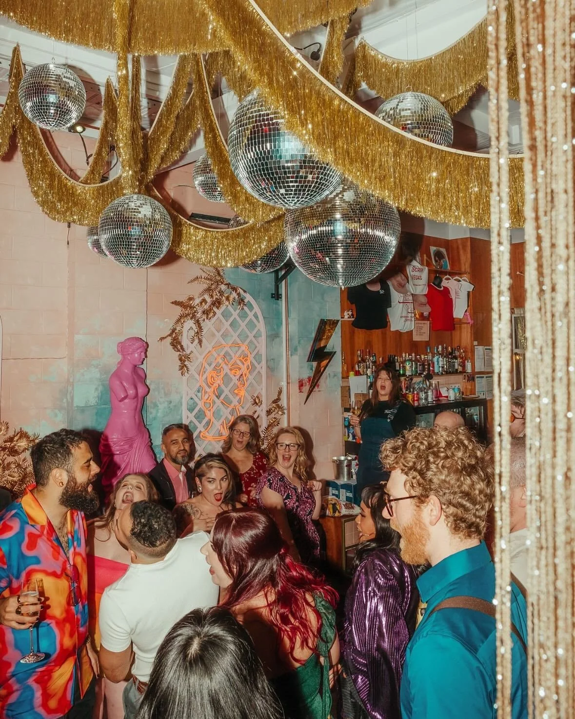 A lively party scene with people socializing and celebrating in a decorated venue featuring disco balls, gold fringe garlands, and a pink statue. The atmosphere is festive with colorful attire and bright lighting.