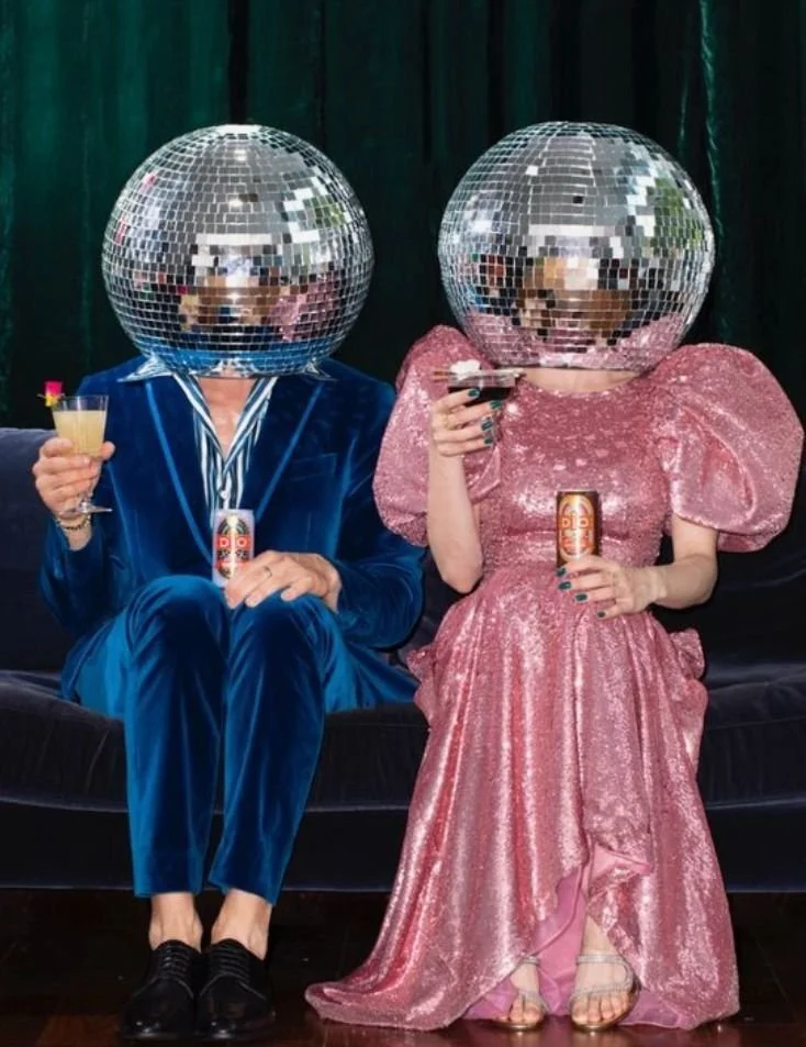 Two people sitting on a black sofa wearing shiny disco ball helmets, one in a blue suit and the other in a pink glittery dress. They are holding drinks and beaded cans, with dark curtains in the background.