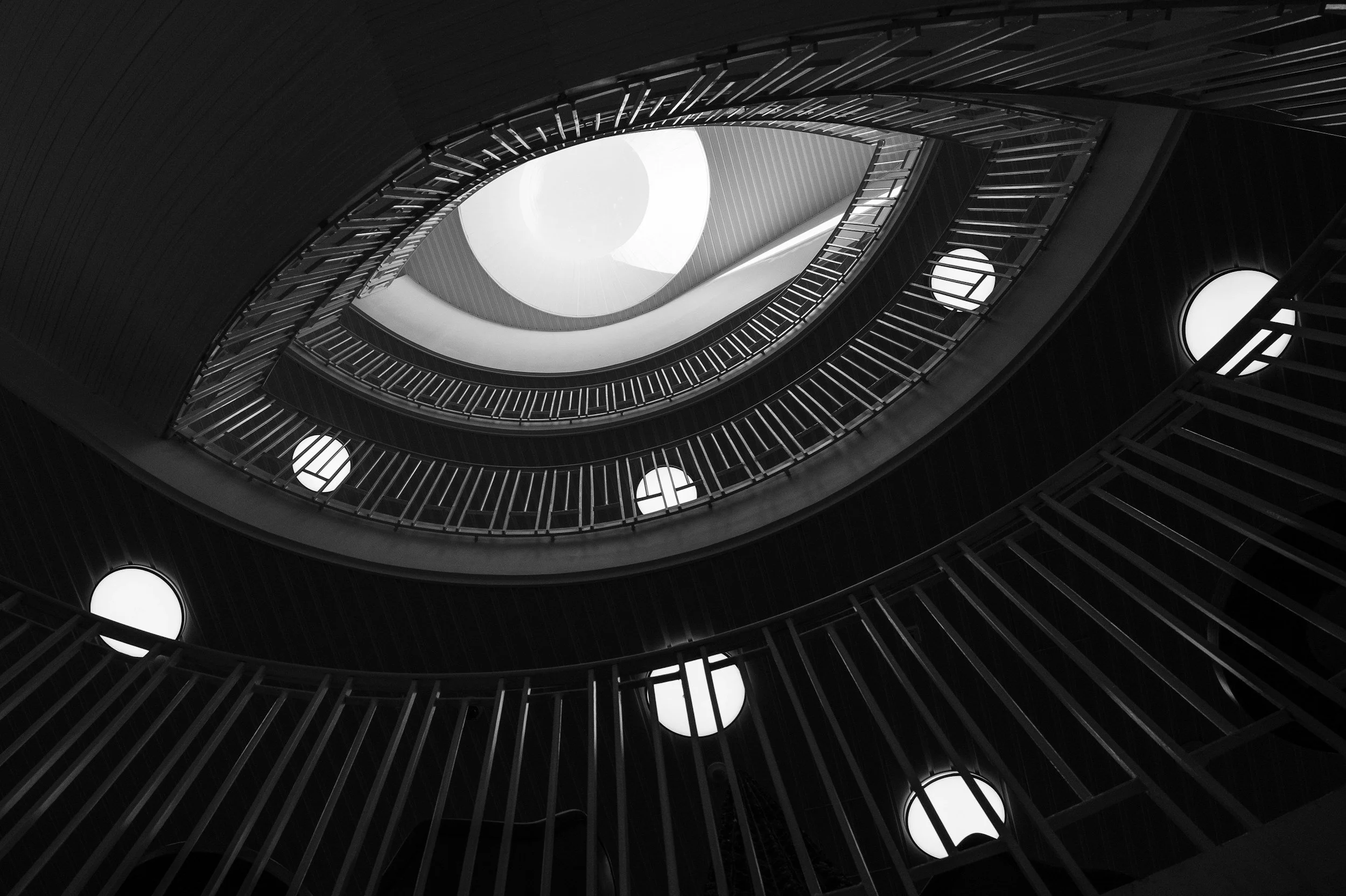 Looking up through a spiral staircase in a tall building, black and white photo, circular windows on the side walls, geometric architectural design.