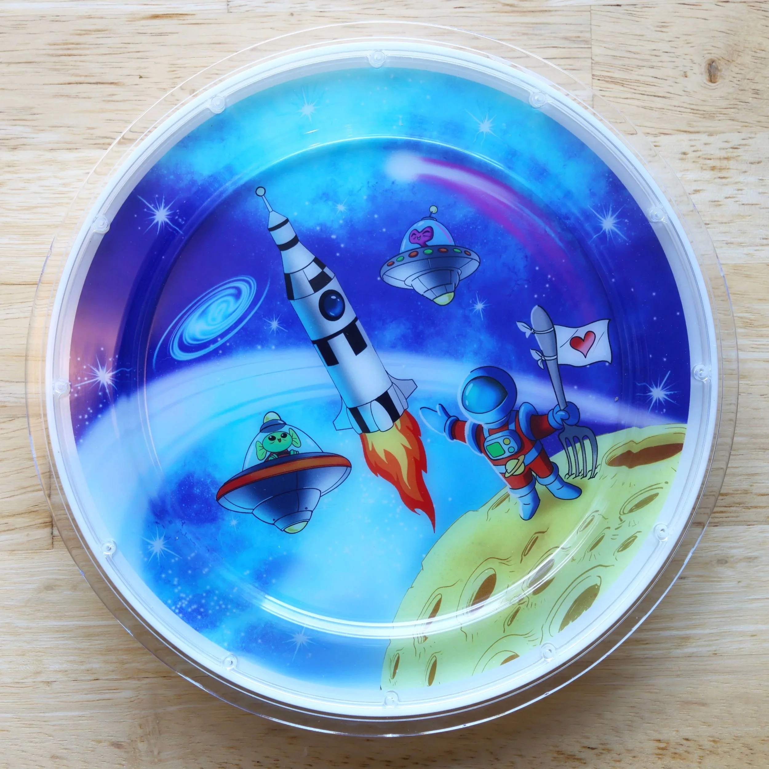 Colorful space-themed illustration on a light up plate, featuring an astronaut with a flag, a rocketship launching, and small alien spaceships with planets and stars in the background.
