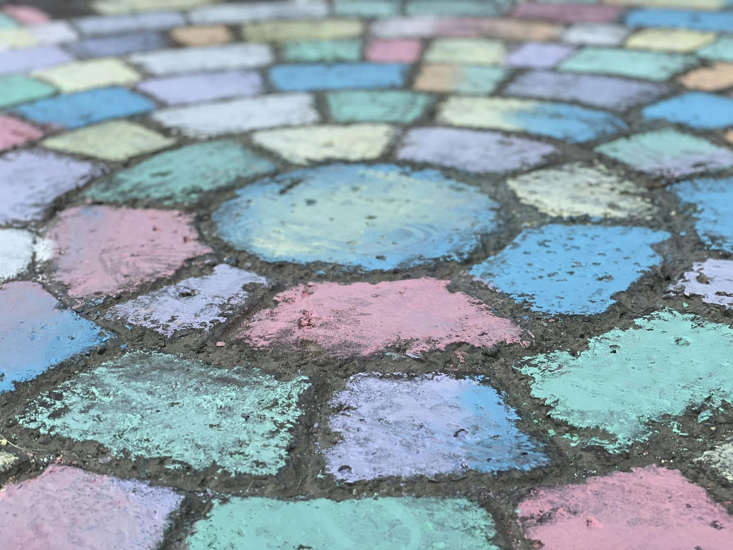 Close-up of a sidewalk with colorful chalk drawings in pink, blue, purple, and green, arranged in a circular pattern.