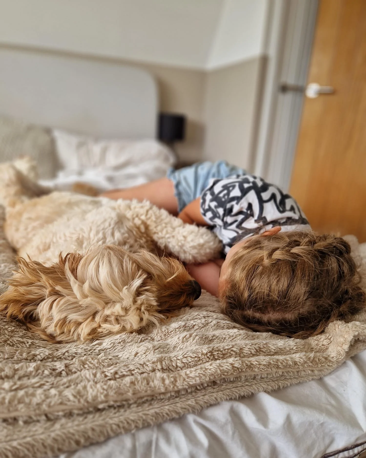 A young child and a small dog sharing a nap on a bed, cuddled together on a soft blanket.