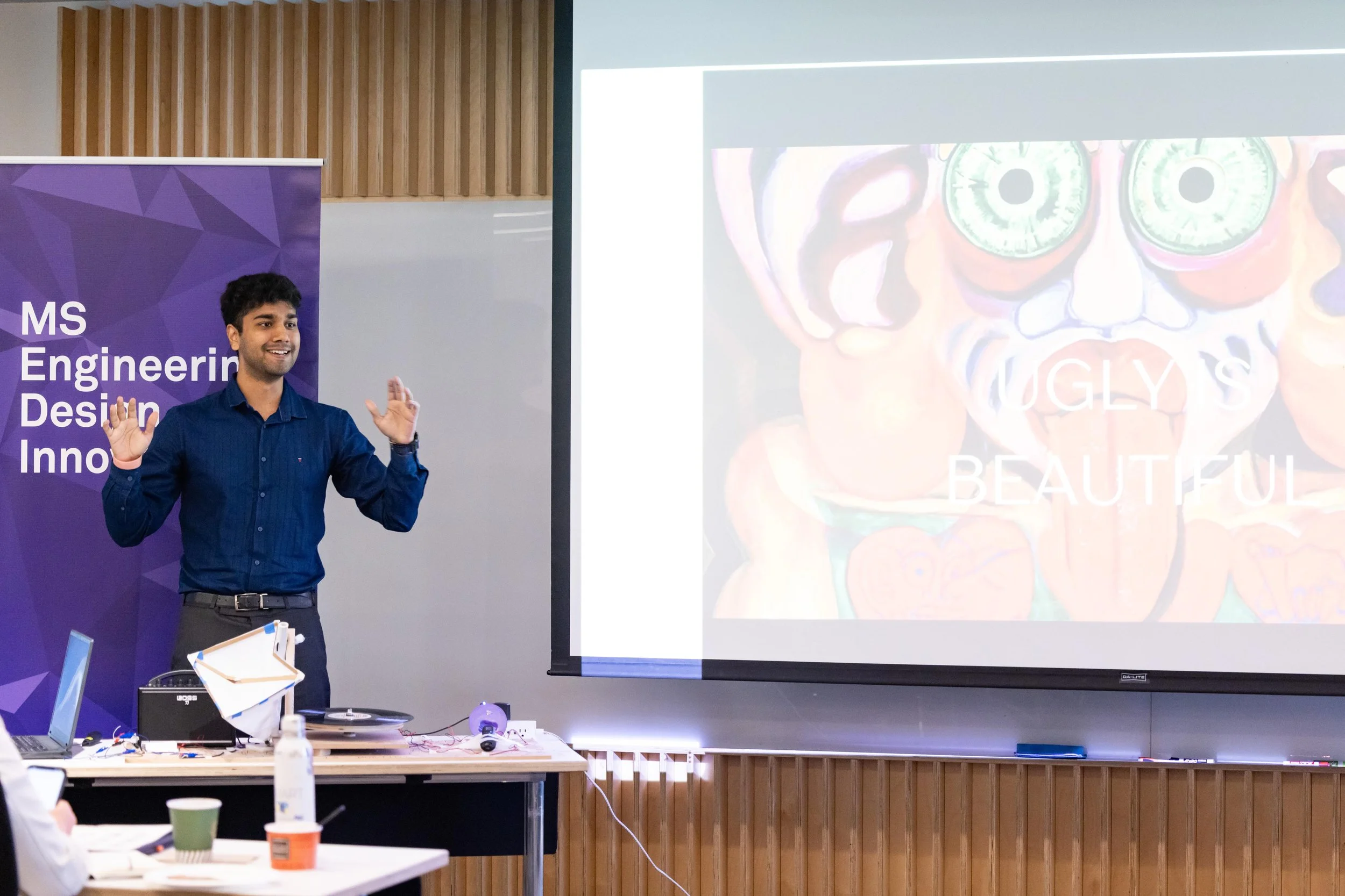 Young man giving a presentation in a classroom or conference room, on a screen behind him a colorful artistic image with the words "UGLY & BEAUTIFUL."