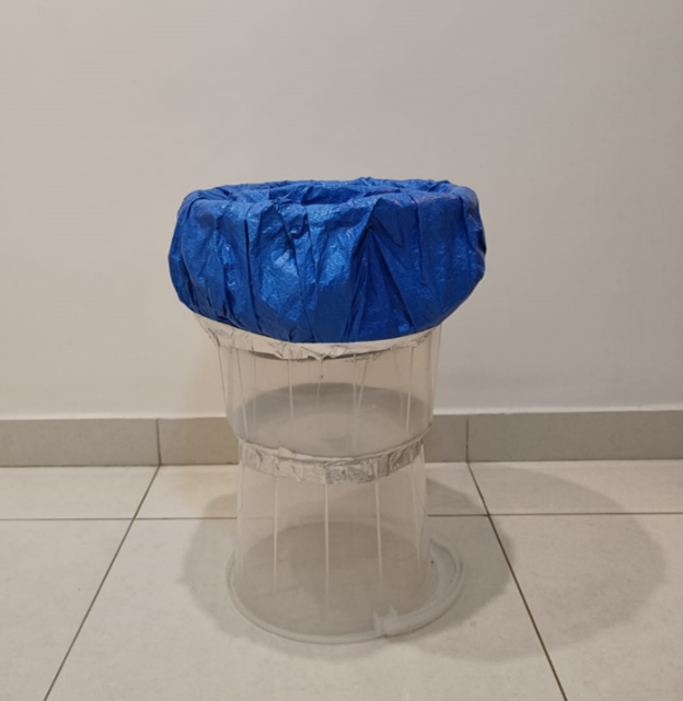 A transparent plastic container with a blue plastic bag covering its opening, placed on a tiled floor against a plain white wall.