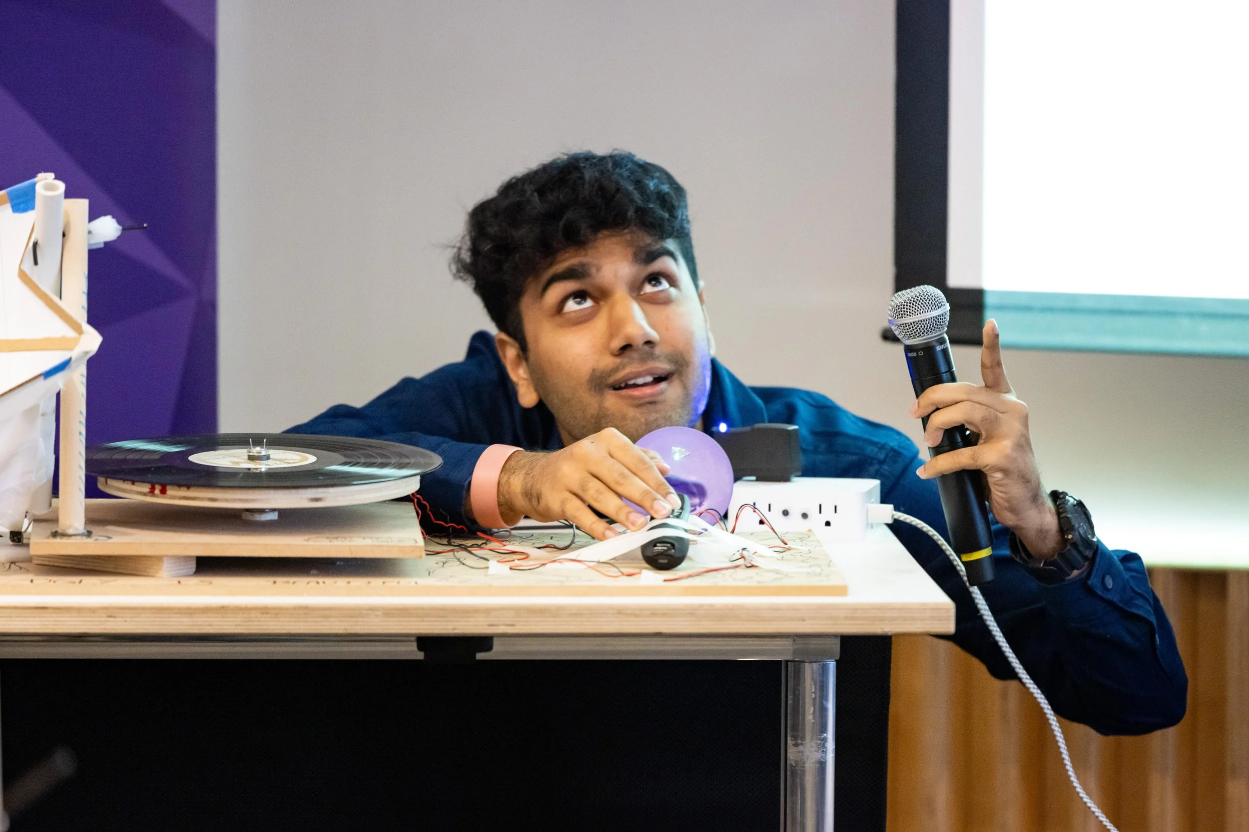 A young man with dark, curly hair, holding a microphone in one hand and touching a light bulb with the other, sitting behind a table with electronic components and a turntable, in a room with a purple and beige wall, appearing to be presenting or demonstrating an experiment or project.