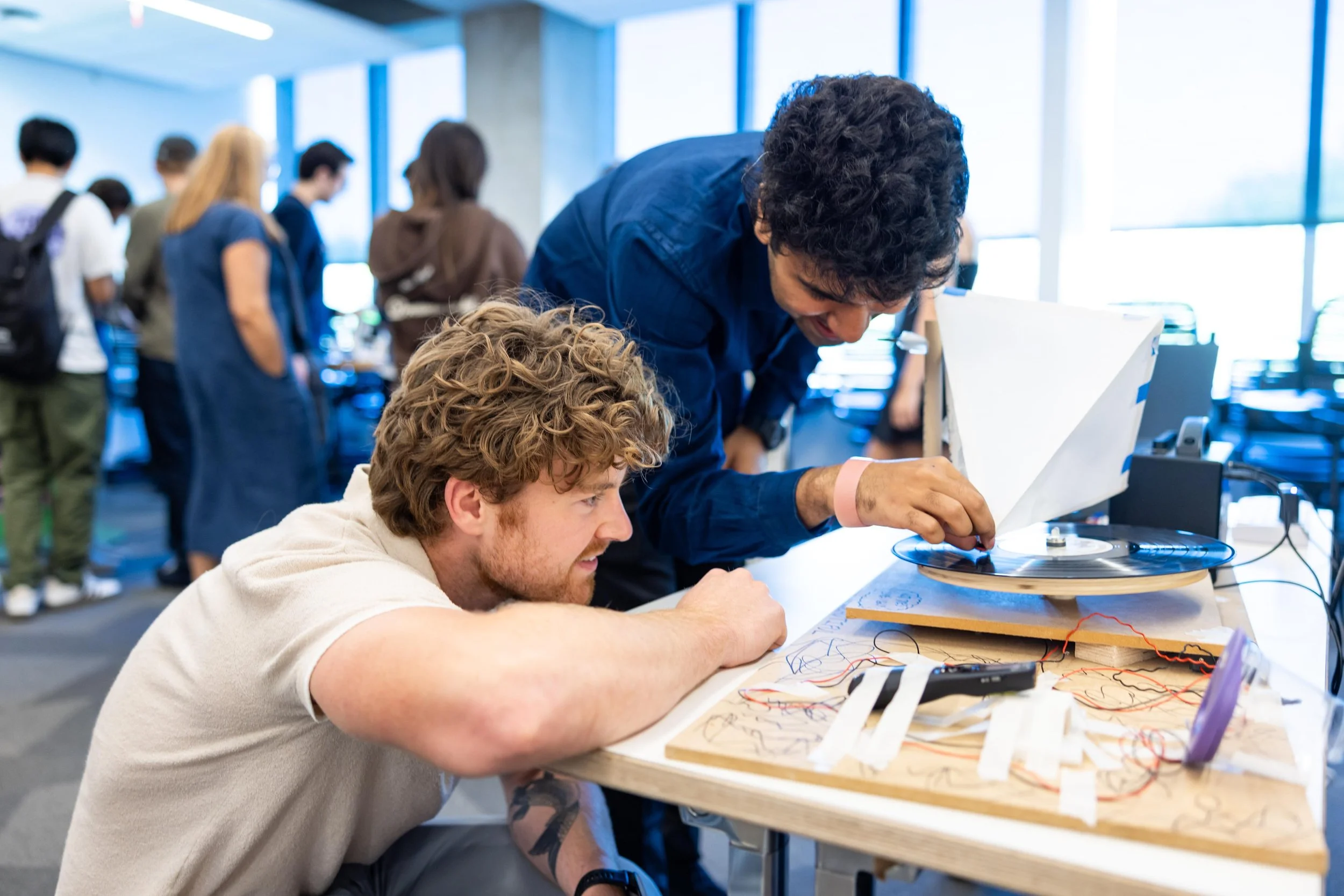 Two young men working together on an electronic project at a desk, with one leaning over and the other sitting, amid a busy room with other people in background.