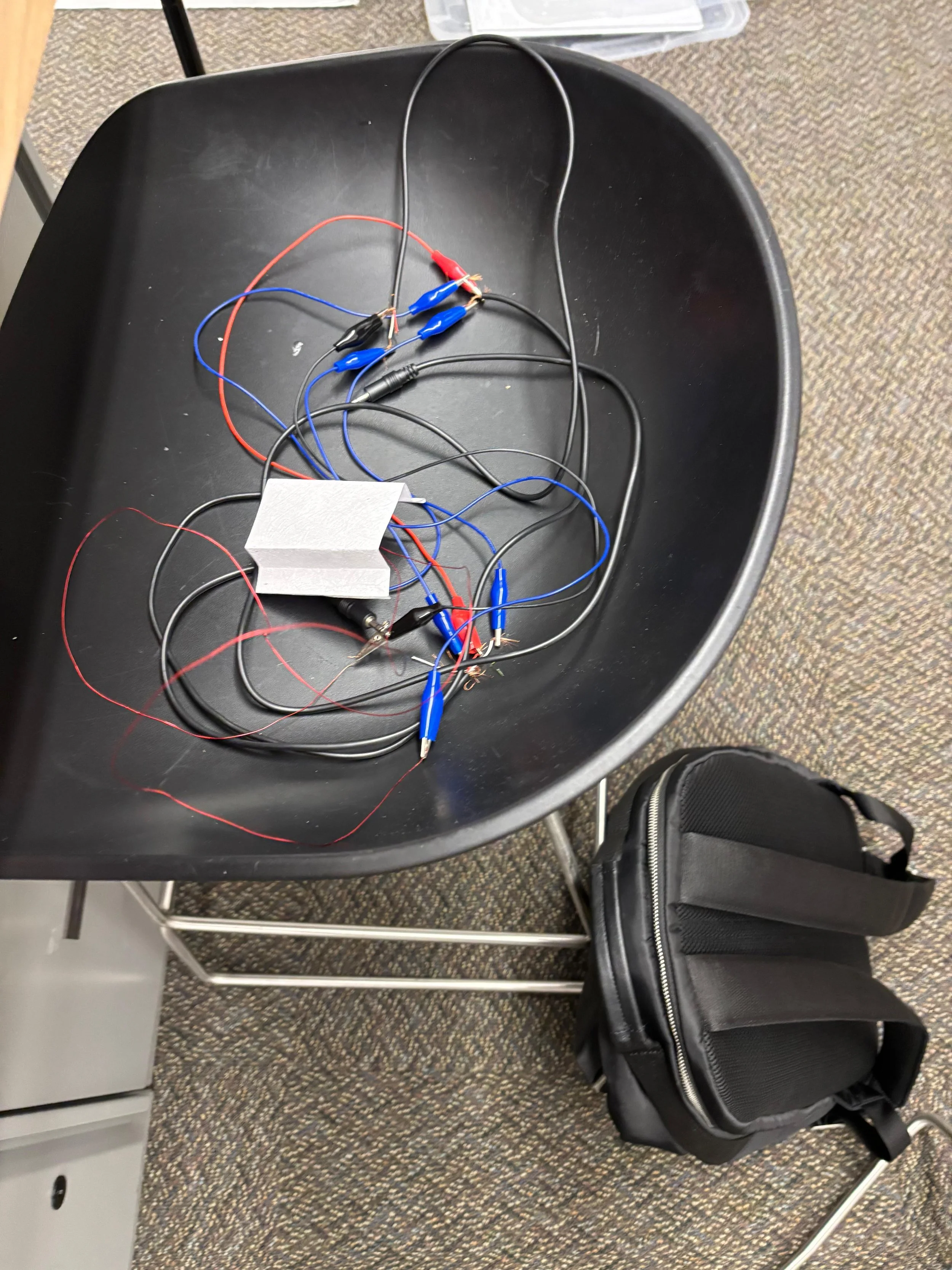 A black tabletop with colorful wires and alligator clips, a foam packet, and a black shoulder bag nearby.
