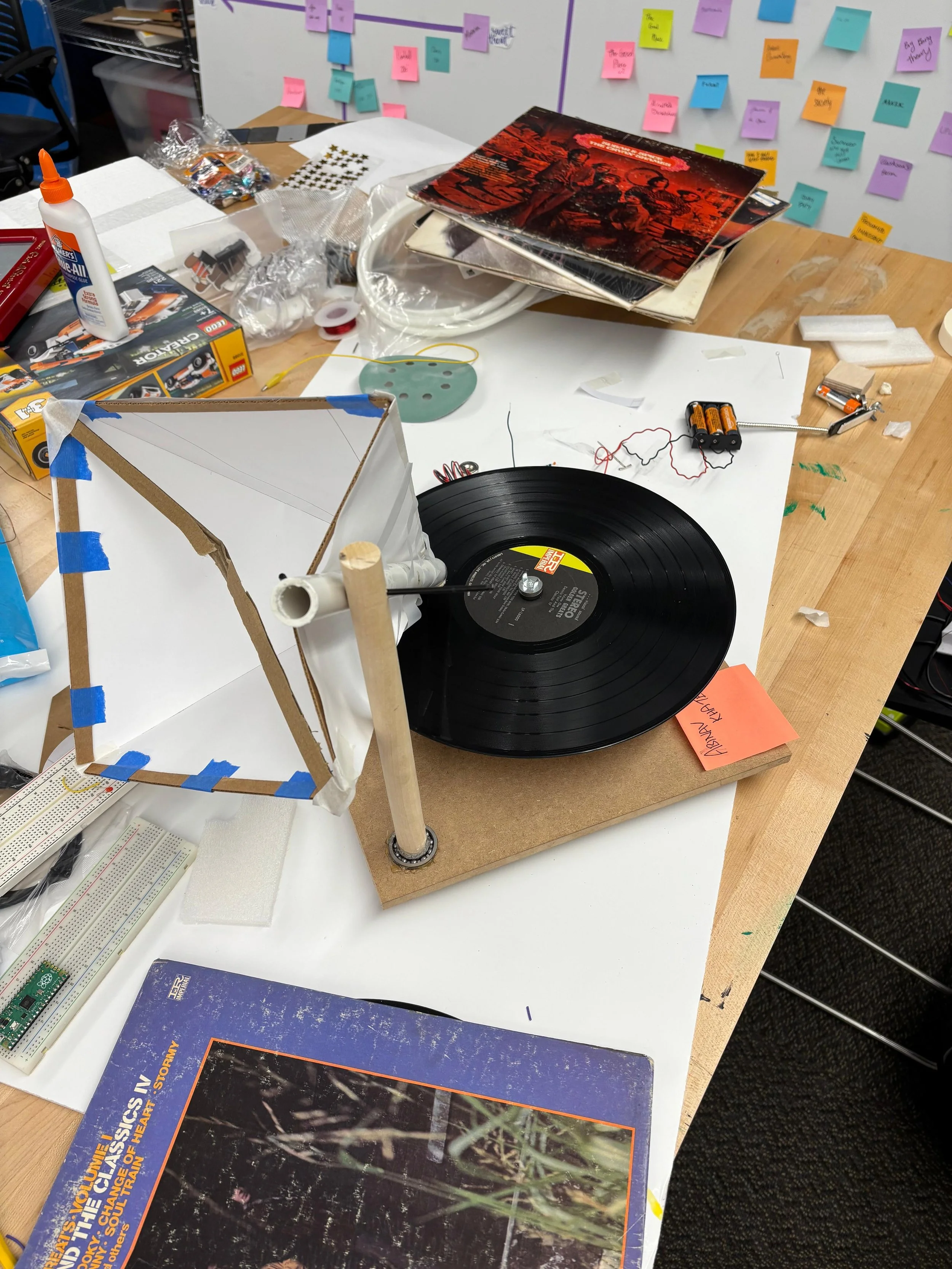 A tabletop space for a science project with a turntable featuring a vinyl record, a makeshift satellite model with a white fabric dish, various electronics, tools, and supplies scattered around, including a glue bottle, batteries, and posters.
