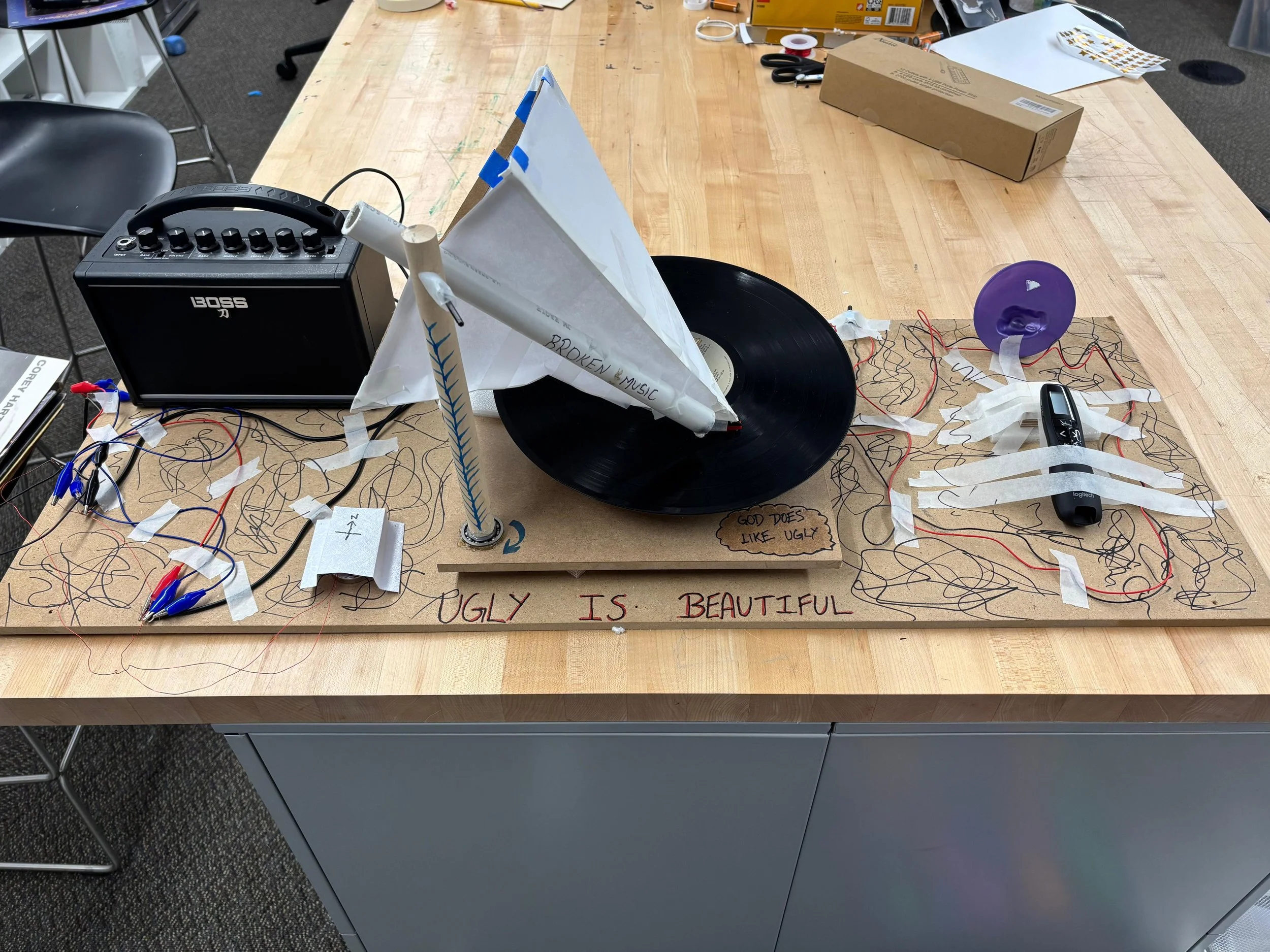 A wooden table holds a messy art project with a vintage vinyl record mounted on a paper cone, a small speaker, wires, a purple plastic cup, a digital thermometer, and a handwritten message on paper reading 'UGLY IS BEAUTIFUL' with additional notes and scribbles.
