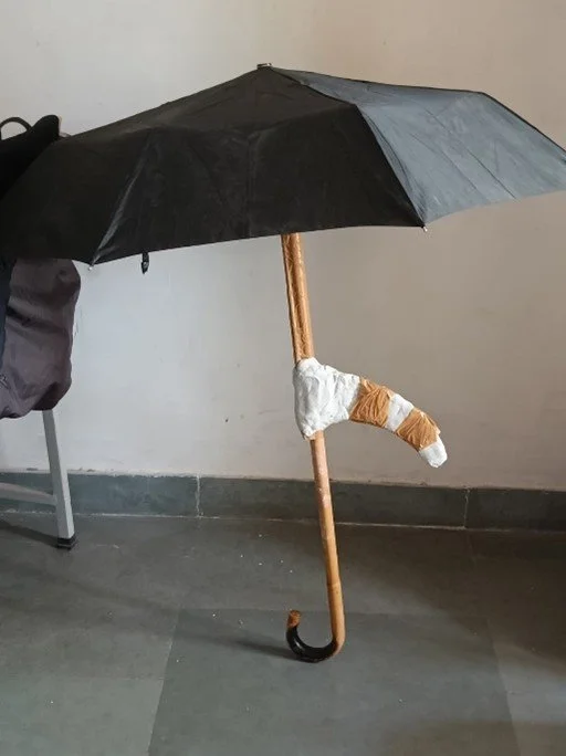 An umbrella with a makeshift arm made of cloth and tape attached to the handle, leaning against a wall.