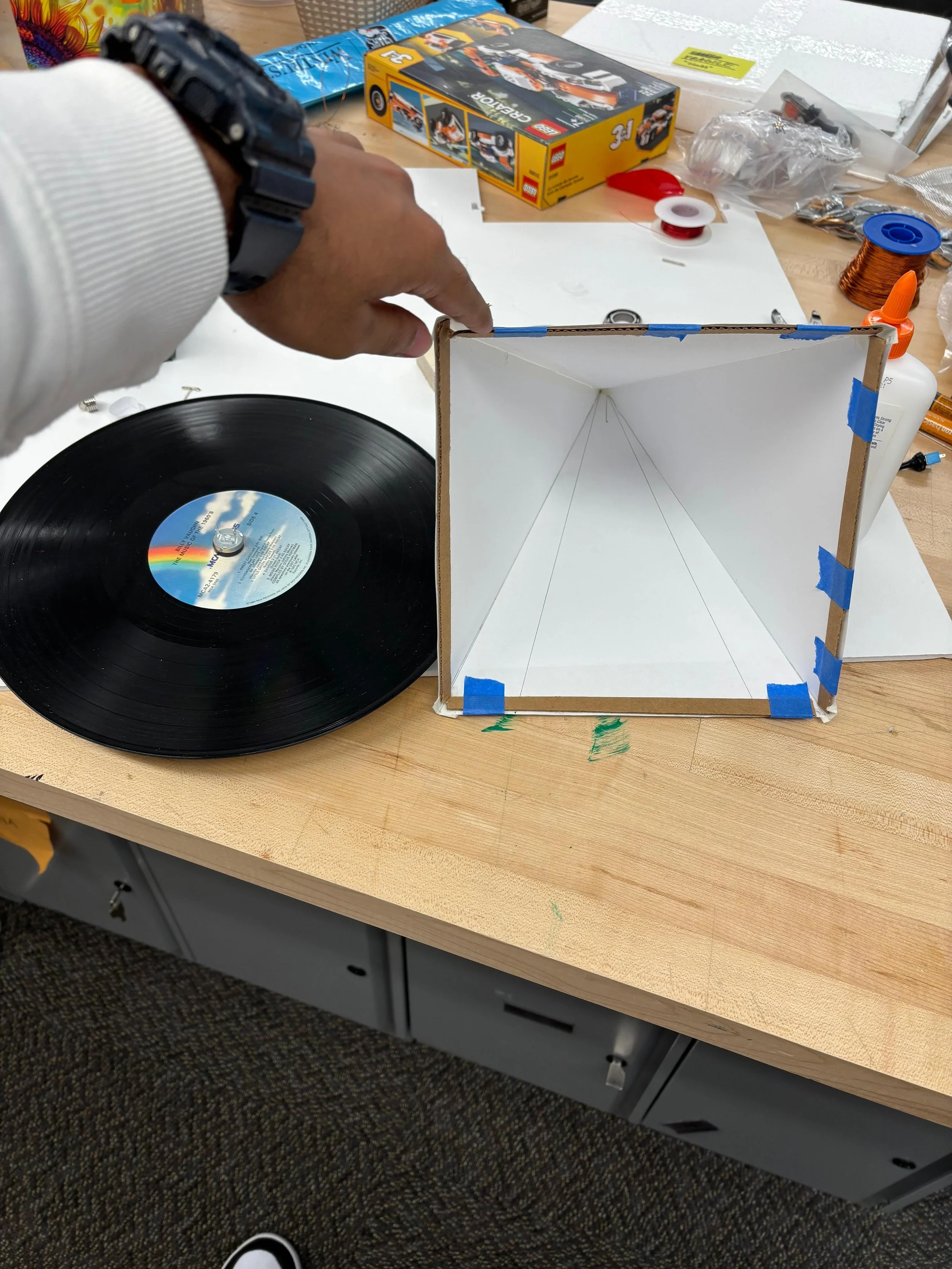 A person assembling a homemade speaker enclosure using a cardboard box, a vinyl record, and various tools.