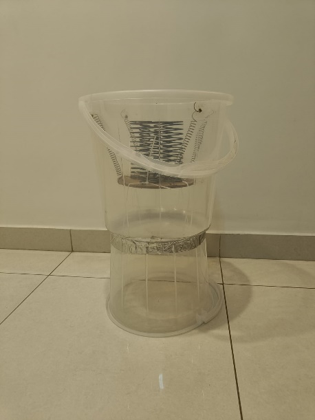 A transparent plastic chair stacked on top of another chair, with the top chair's seat inclined and a coiled spring visible inside it, placed against a plain wall on tiled flooring.