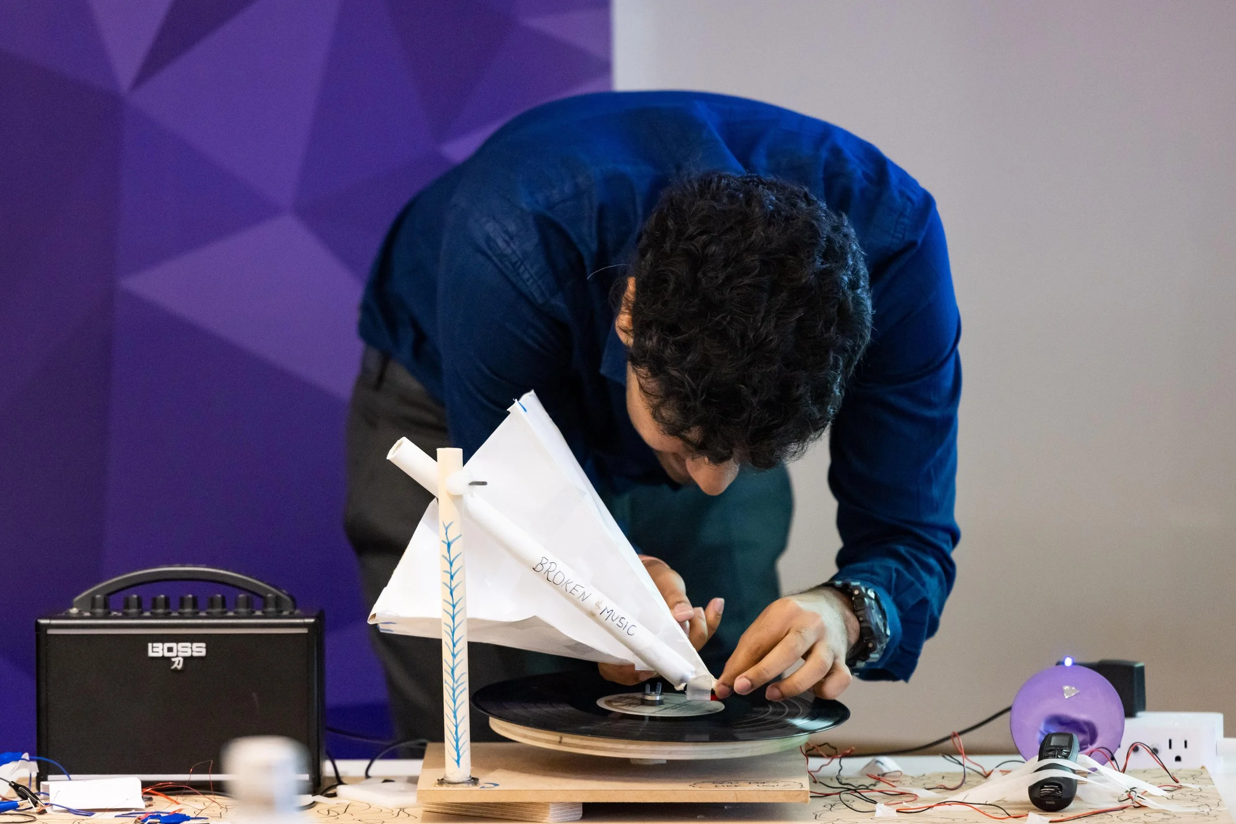 A person with curly hair wearing a dark blue shirt leans over a table, adjusting a small homemade wind turbine with a fabric sail marked 'Broken Music' attached. The table has electronic components, a small amplifier, wires, a purple speaker, and a vinyl record.
