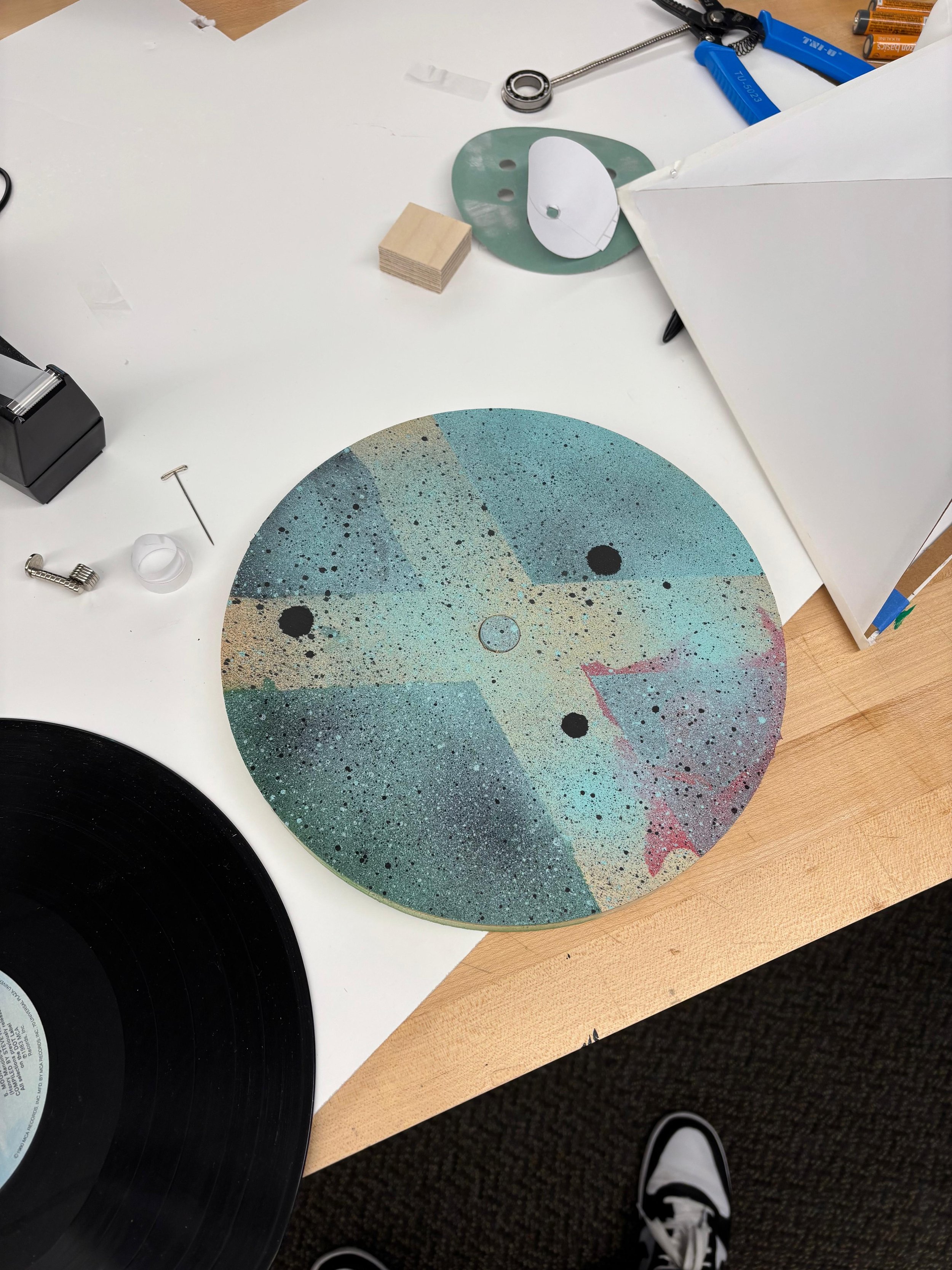 A colorful, speckled circular object with black dots on a white table, surrounded by various tools and objects including a vinyl record, a white lightbox, a green stencil, and small wooden blocks.