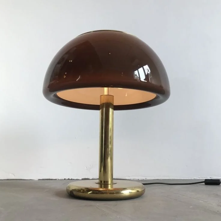 Luminária de mesa moderna com base dourada e abajur marron, ligada em uma parede branca.
