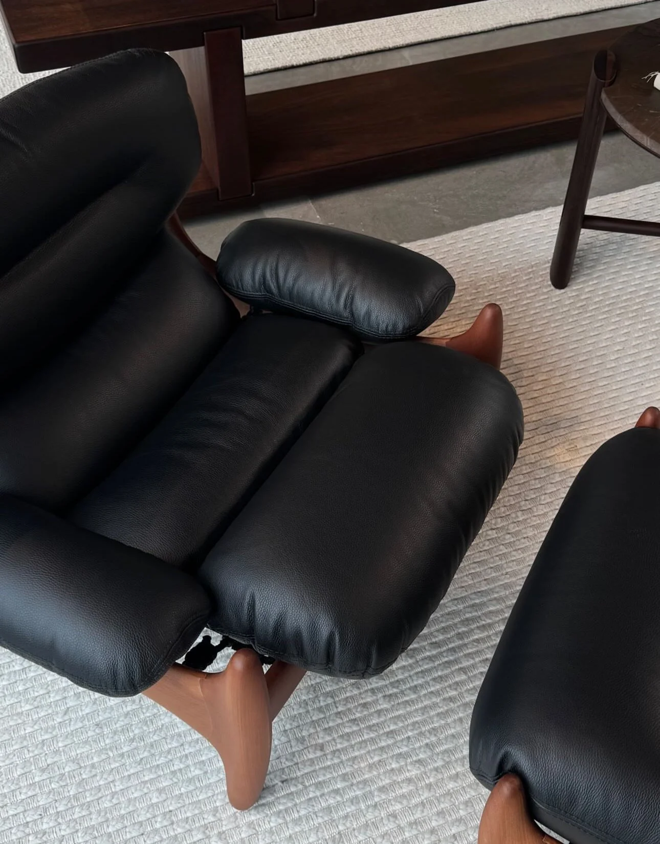 Sofá de couro preto com estrutura de madeira, em um ambiente com tapete branco e móveis de madeira ao fundo.