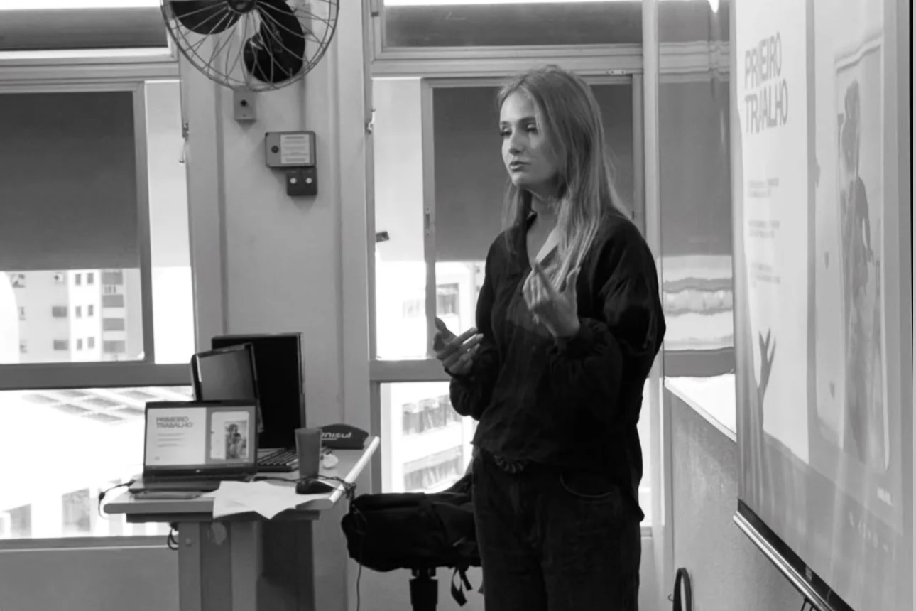 Mulher dando uma apresentação em uma sala de aula ou reunião, ao lado de um quadro e uma mesa com computador e livros.