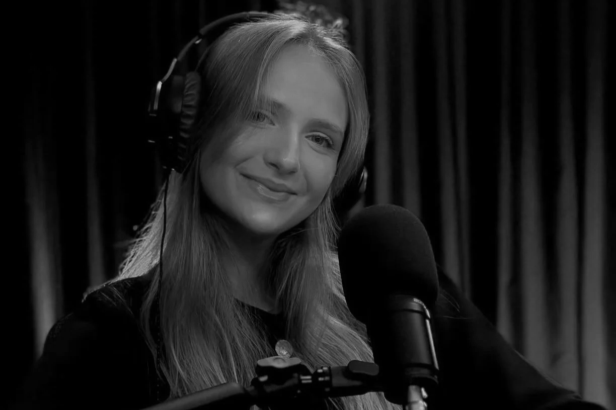 Mulher sorridente com cabelo loiro, usando fones de ouvido sobre uma camisa escura, falando numa gravação de áudio ou rádio, com fundo de cortinas longas.