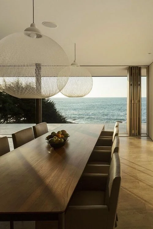 Sala de jantar com mesa de madeira e cadeiras ao lado de uma janela grande com vista para o mar.