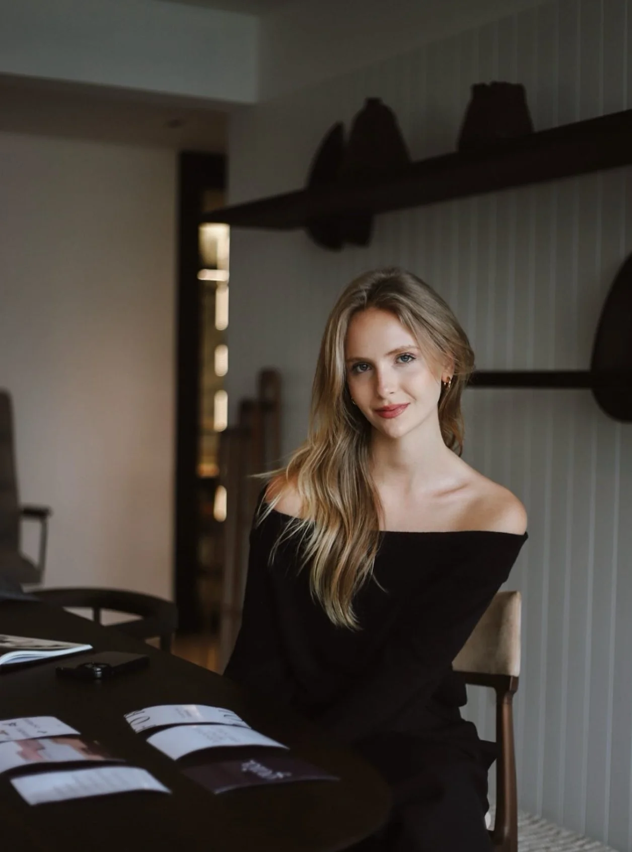 Mulher de cabelo loiro e cabelo ondulado, usando blusa preta, sentada em uma mesa com revistas e telefone, dentro de uma sala com paredes com painéis de madeira.