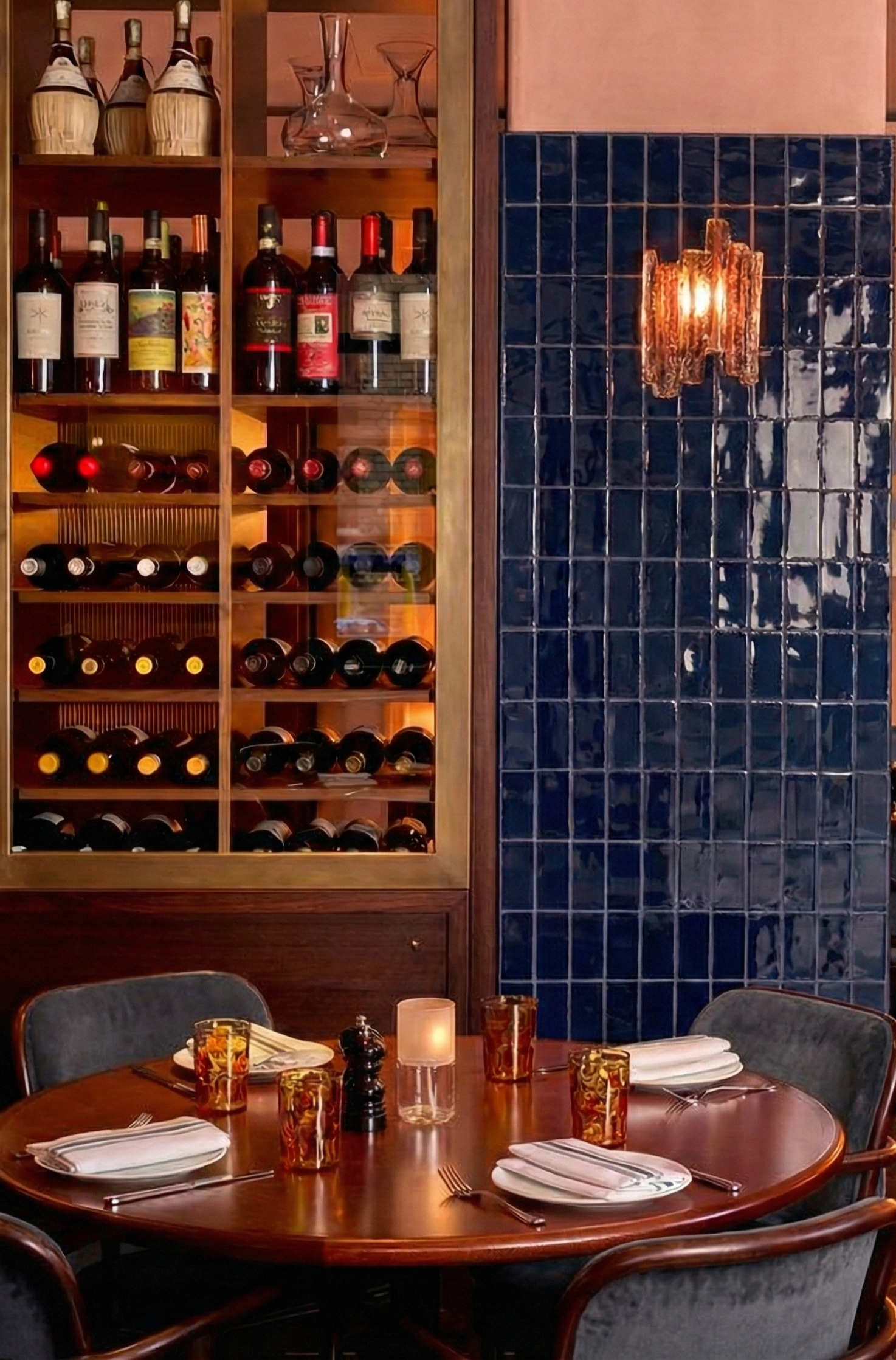 Sala de jantar com mesa de madeira, quatro talheres, copos ambar, pratos brancos e velas, parede de azulejos azuis e prateleira com garrafas de vinho e taças.