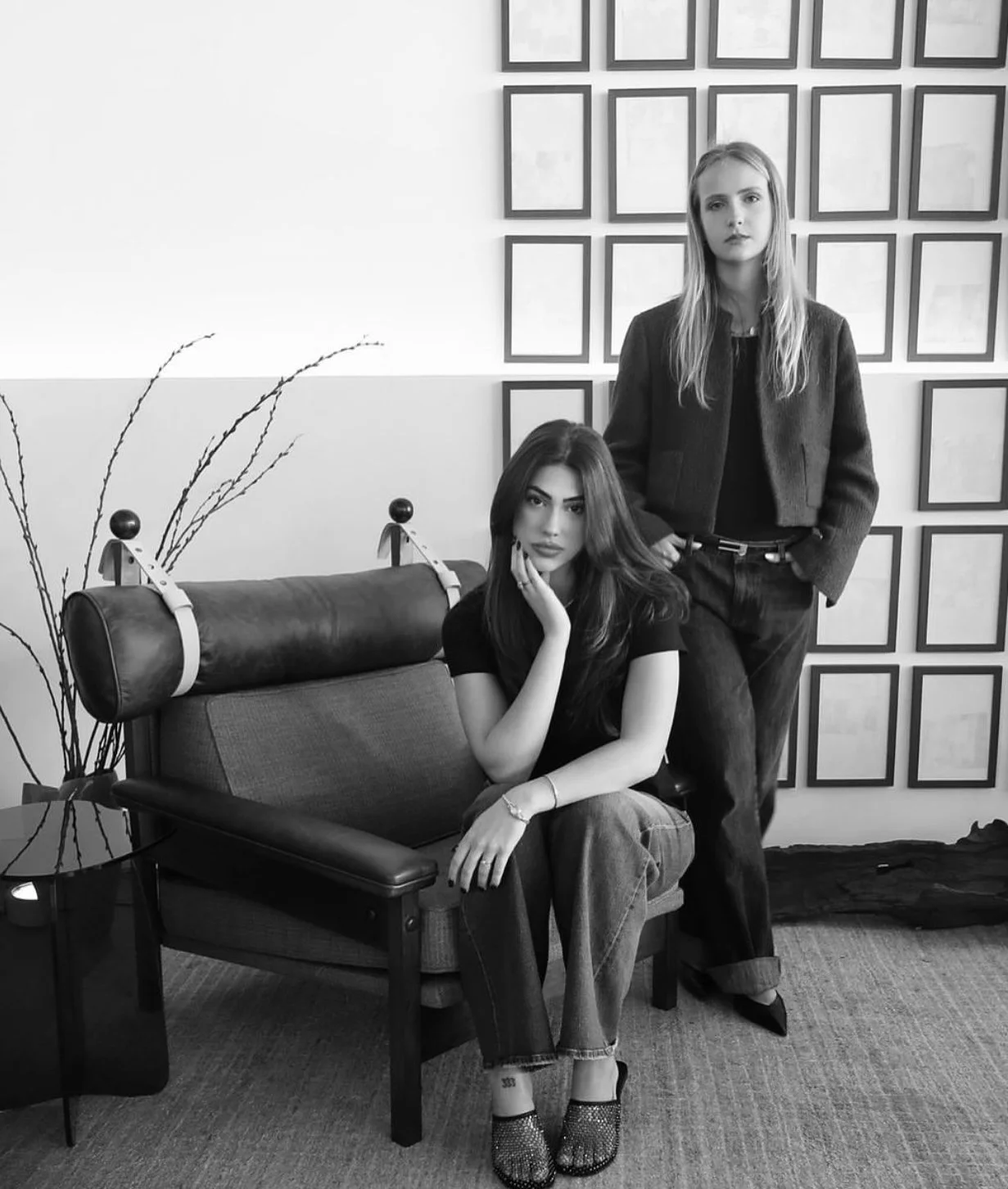 Duas mulheres em um ambiente de sala de estar, uma sentada no sofá com cabelo longo e vestida de preto, a outra de pé ao lado, com cabelo longo e vestido escuro, parede decorada com quadros vazios ao fundo.