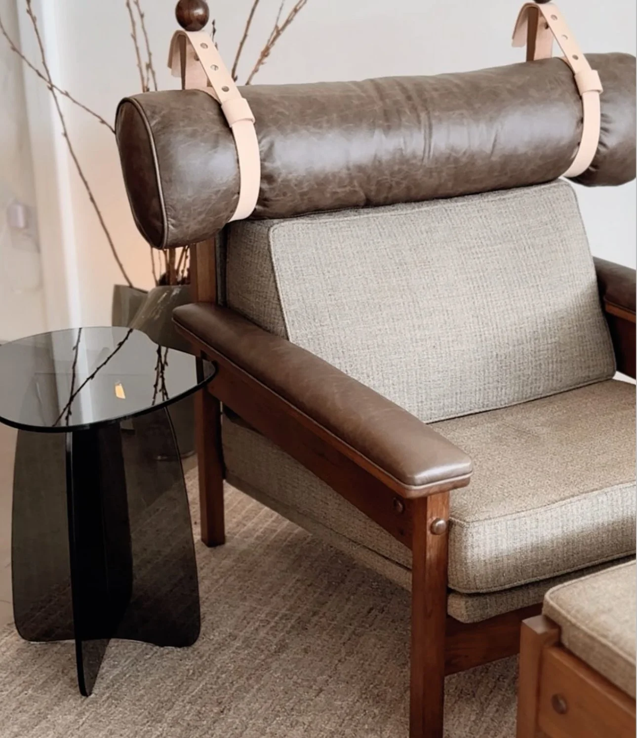 Sofá de madeira com almofadas de estofado bege e encosto de couro marrom. Ao lado, mesa de canto de vidro escuro com pé de suporte preto.